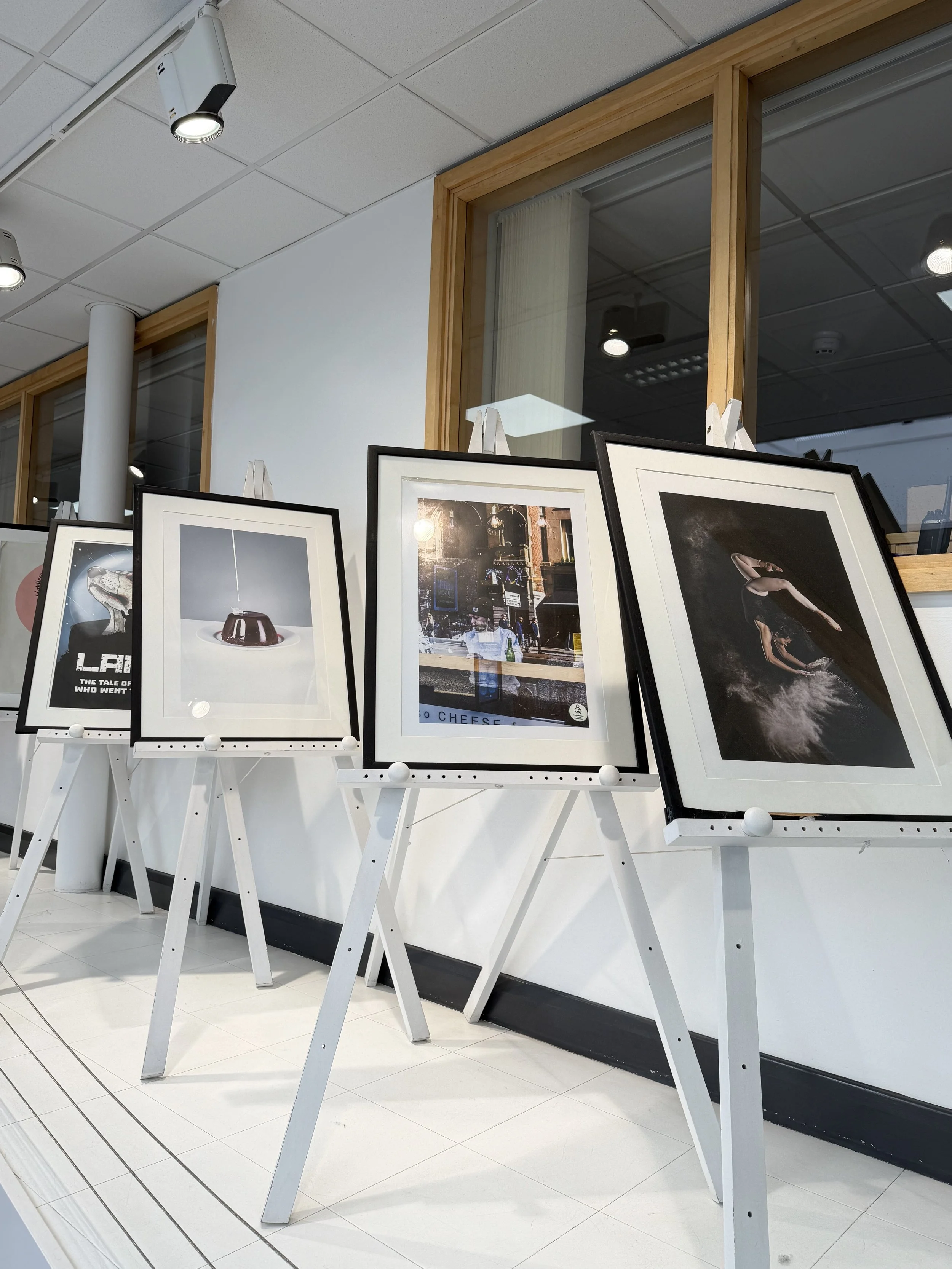 Display of framed photographs on white easels in a modern gallery with white tiled floor and ceiling, wooden framed window, and ceiling spotlights.