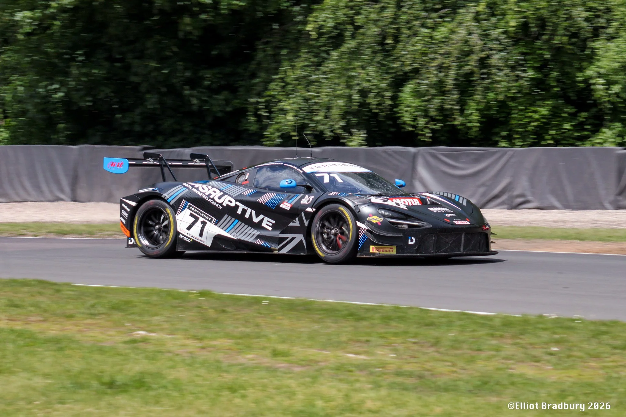 A black race car with blue and white decals driving on a race track with green trees in the background.