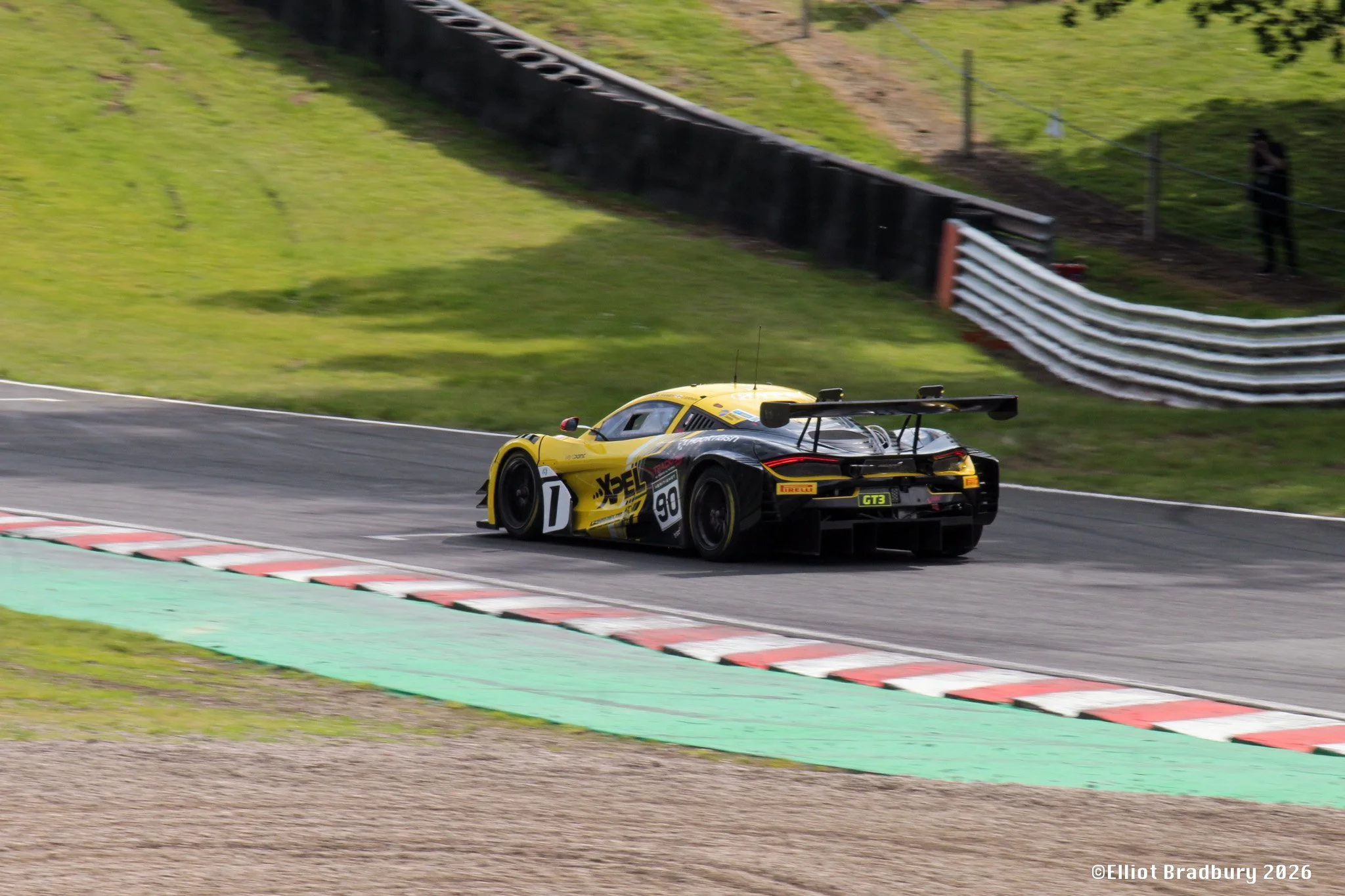 A yellow and black race car speeding on a track with grass and safety barriers in the background.