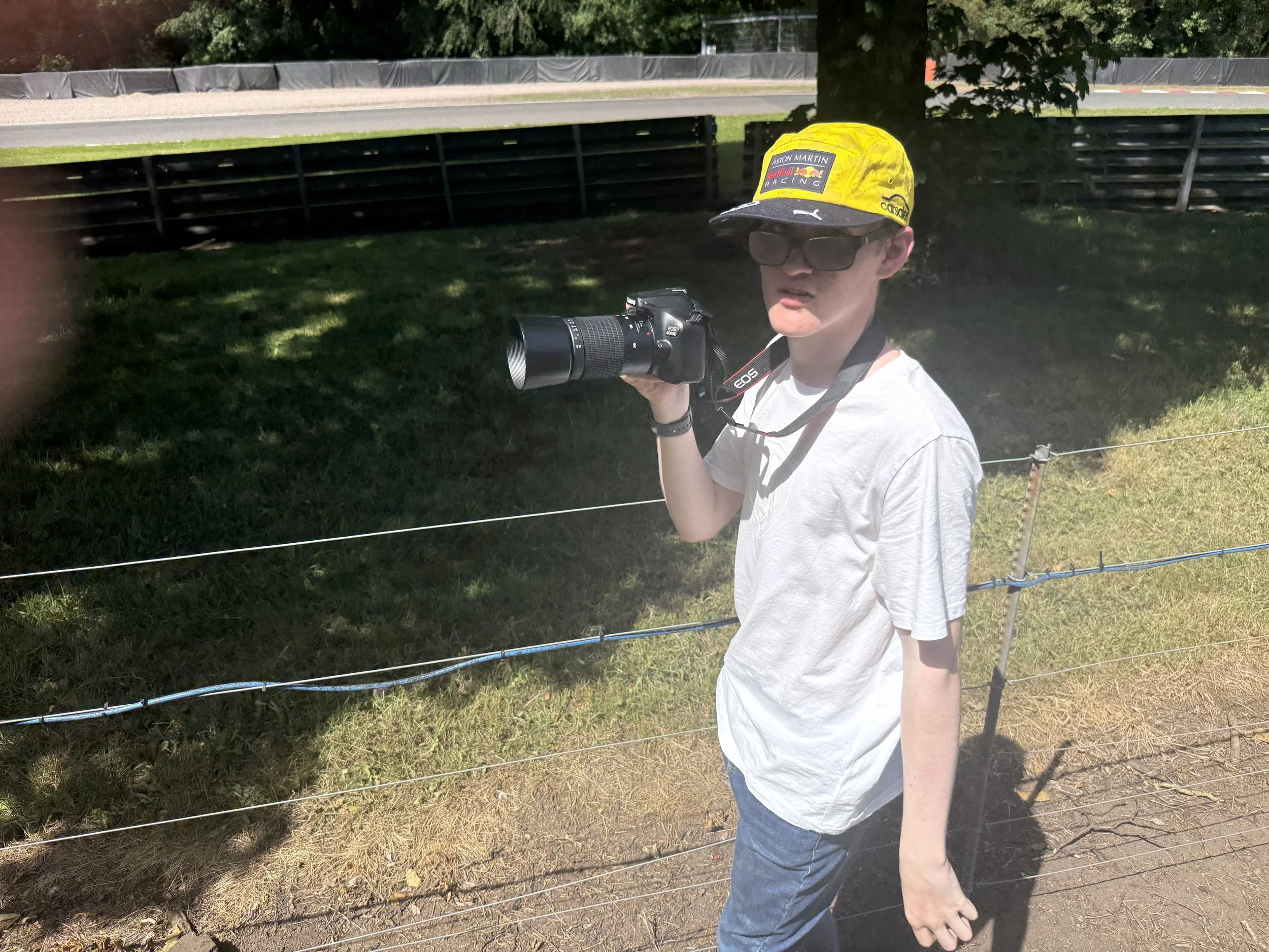 A young man wearing sunglasses and a yellow cap with Racing logos stands outdoors near a fence holding a camera with a telephoto lens.
