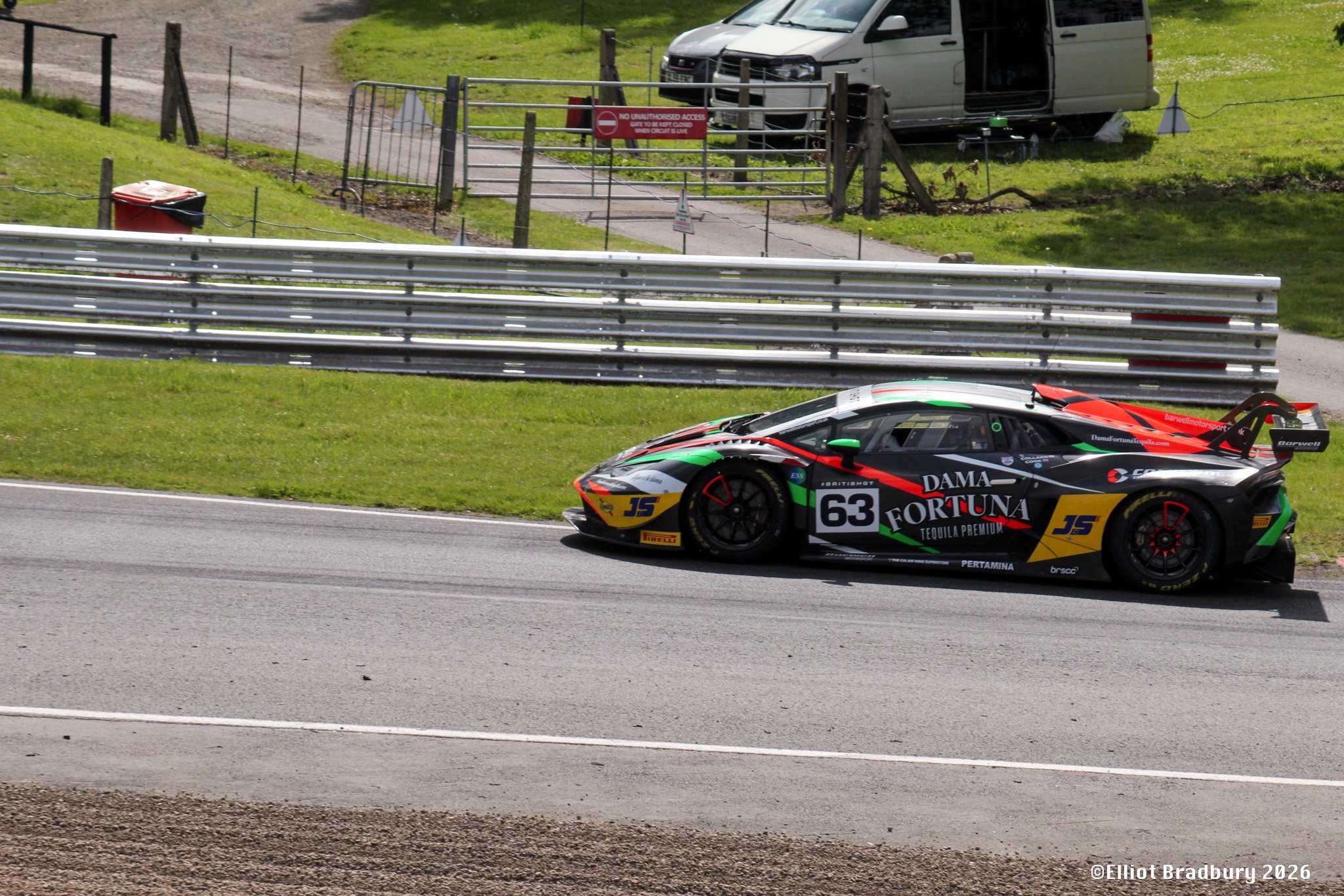 A race car with various sponsor logos moving on a track, with a grassy hillside, a metal barrier, and a gravel area in the background, and a parked van and fencing beyond the barrier.