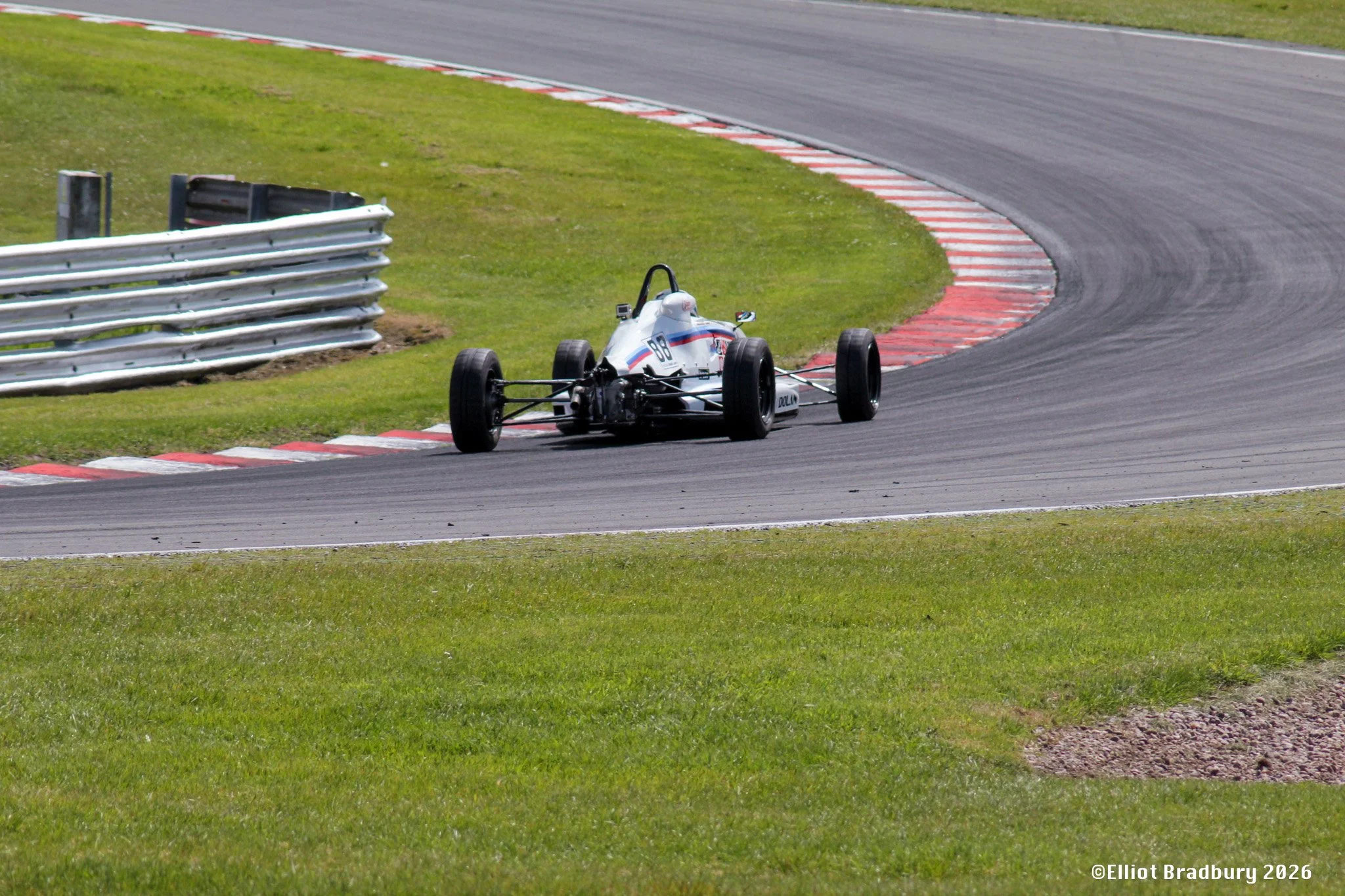 A vintage open-wheel race car on a racetrack, navigating a sharp turn with red and white curbing, surrounded by green grass and a metal guardrail.