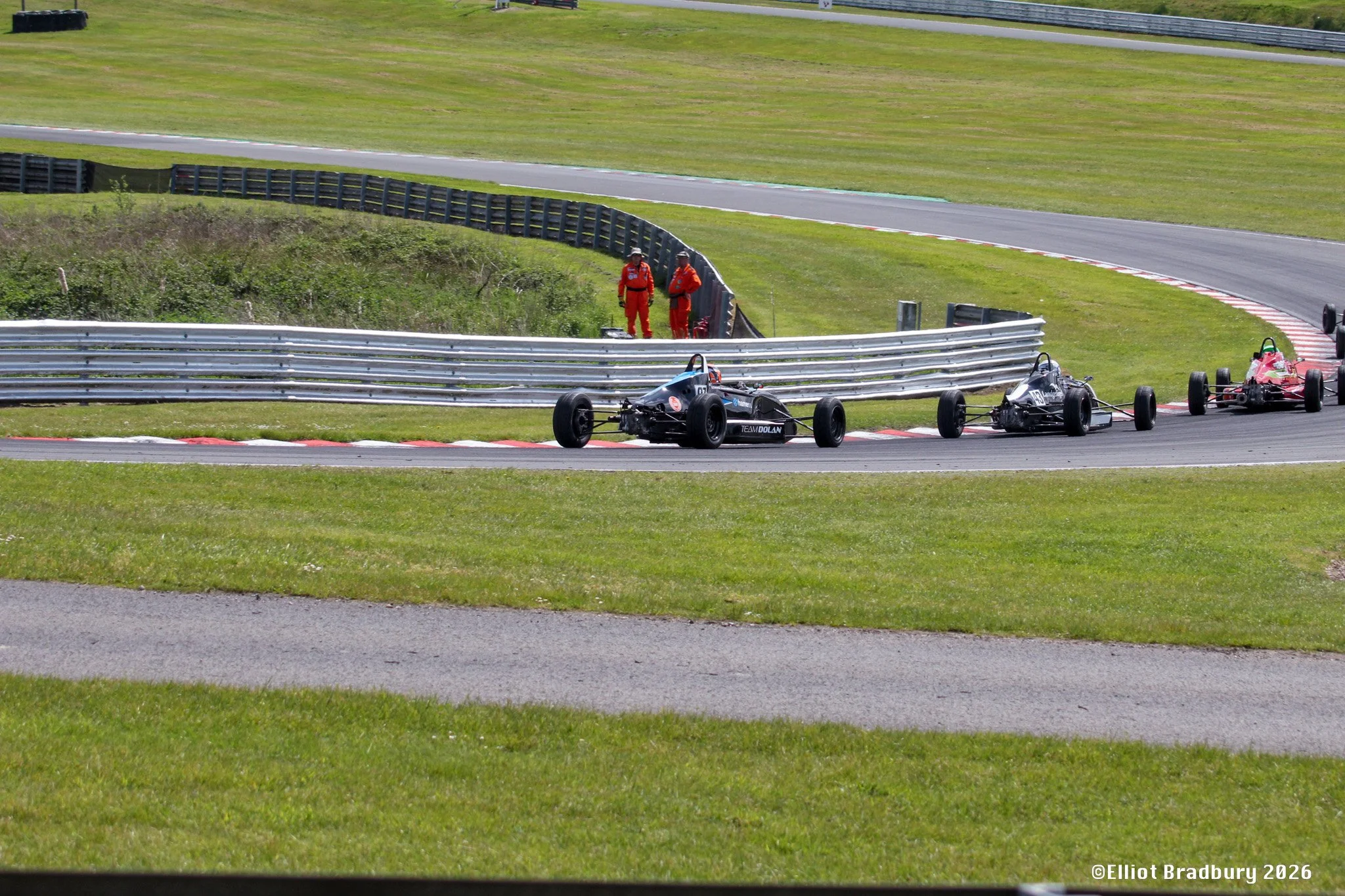 Race cars on a race track with two officials in red jumpsuits standing near the track. The scene is outdoors on a sunny day with green grass and metal guardrails.