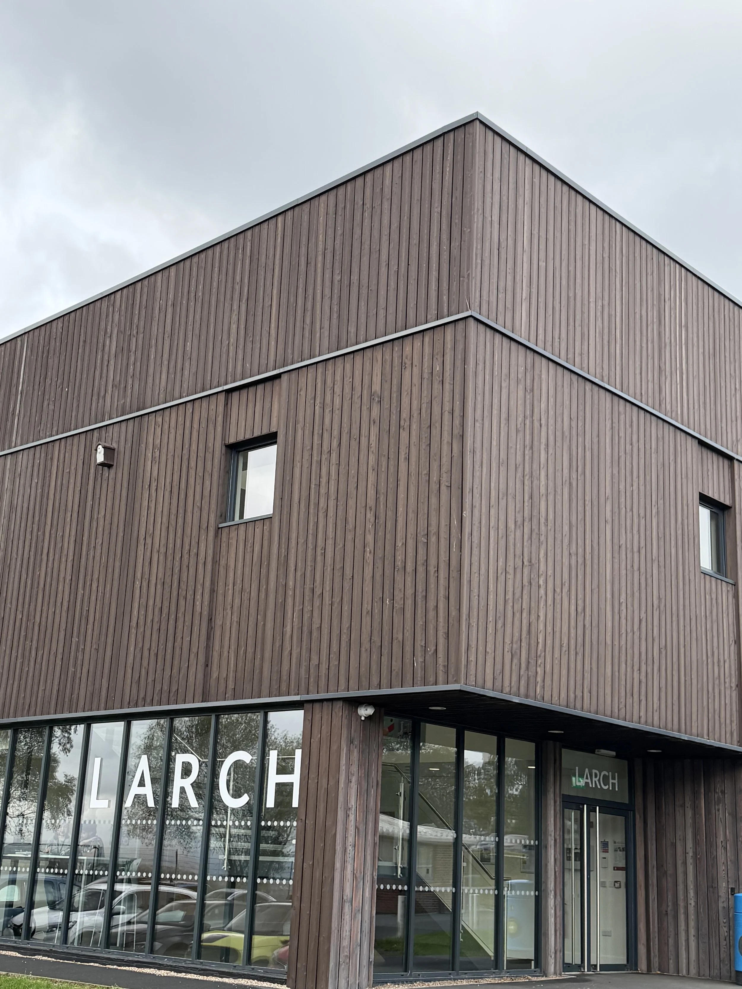 A modern building with a wooden exterior and large glass windows, with the word LARCH on the window, and a cloudy sky above.