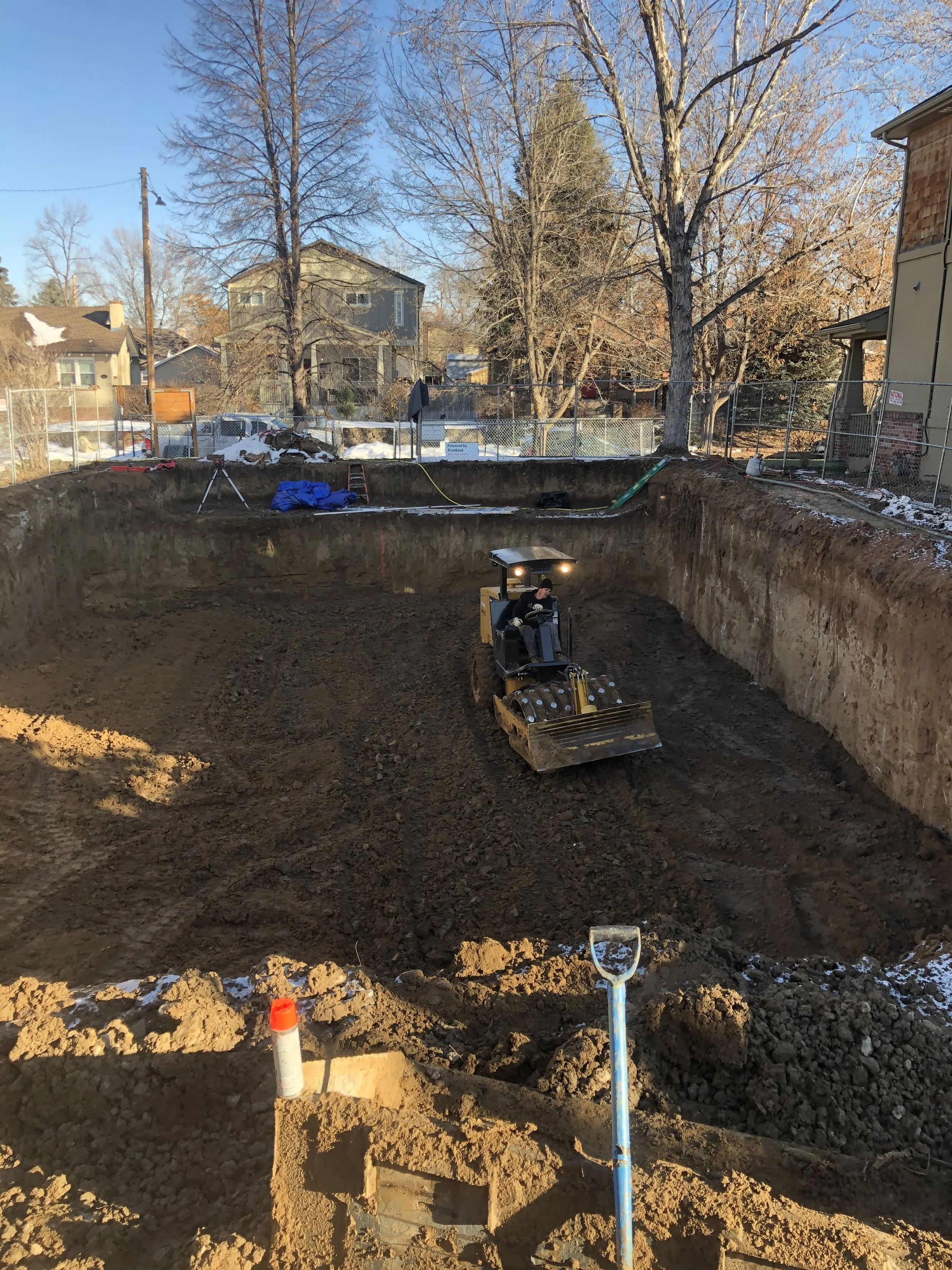Construction site with a yellow bulldozer leveling the dirt in a large excavated area, surrounded by trees and residential houses.