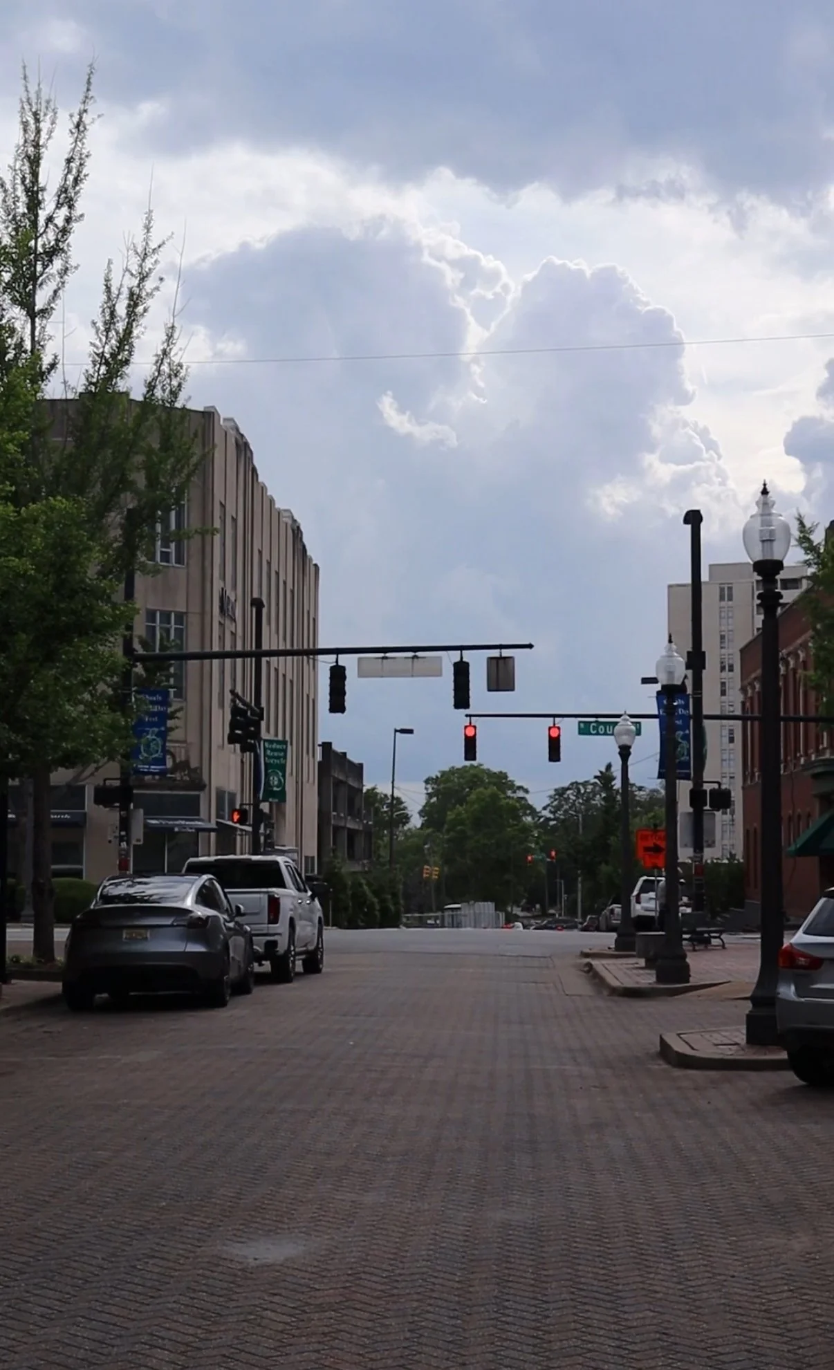 Empty city street intersection under cloudy sky, traffic lights red, parked cars on the curb, streetlamps, buildings, and trees.