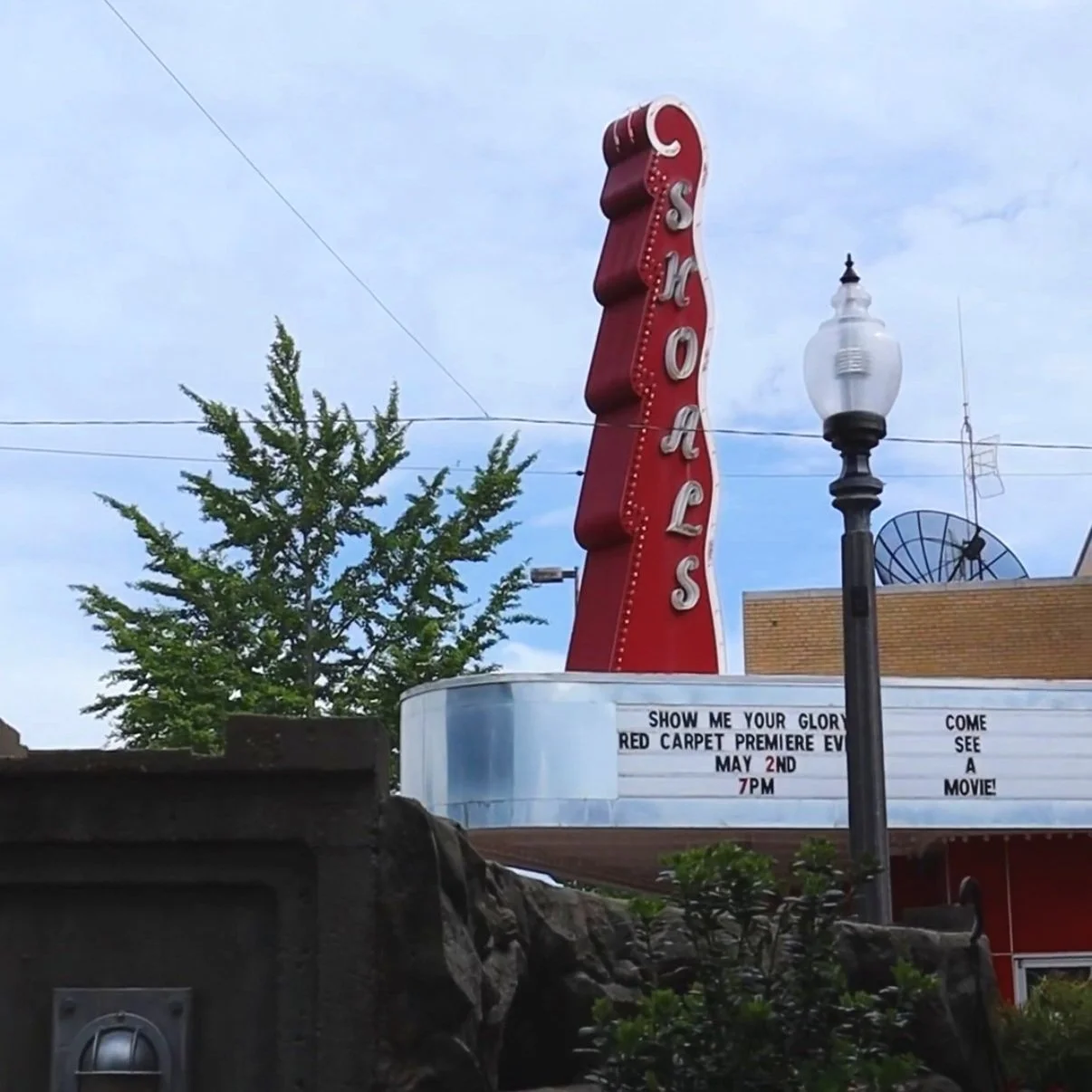 Retro theater marquee with vertical red neon sign reading 'SHOWS'. The marquee displays information about a movie premiere event on May 2nd at 7 pm, with some obscured text and street lamps nearby, under a cloudy sky.