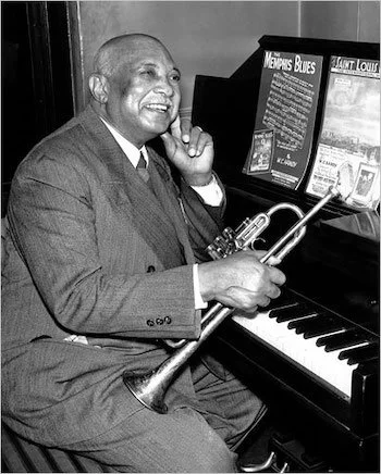 A man in a suit sitting at a piano, holding a trumpet, smiling, with music posters on the wall behind him.