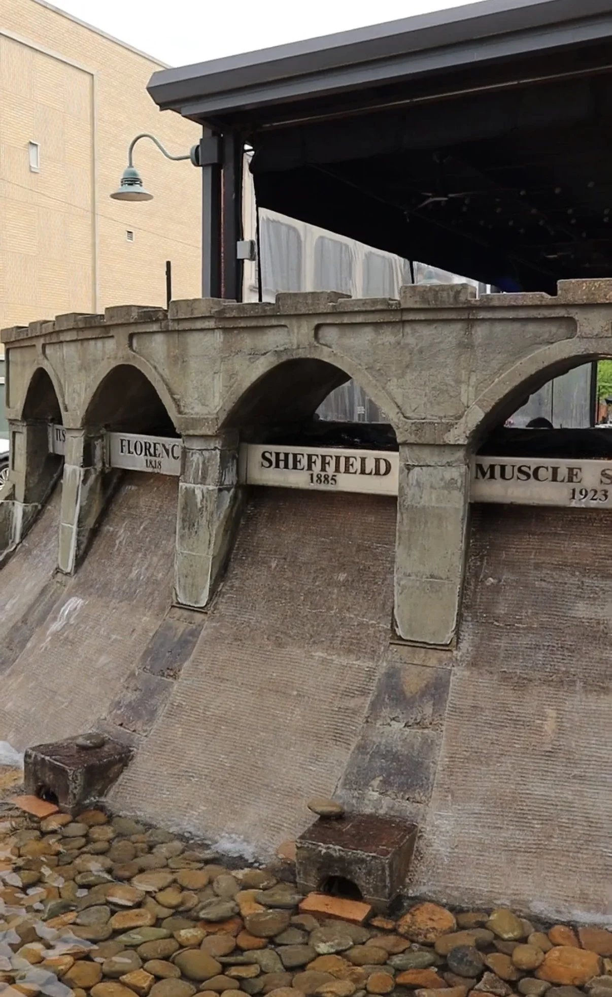 Miniature model of a stone arch bridge with nameplates for different locations and years, displayed over a pebbled ground.