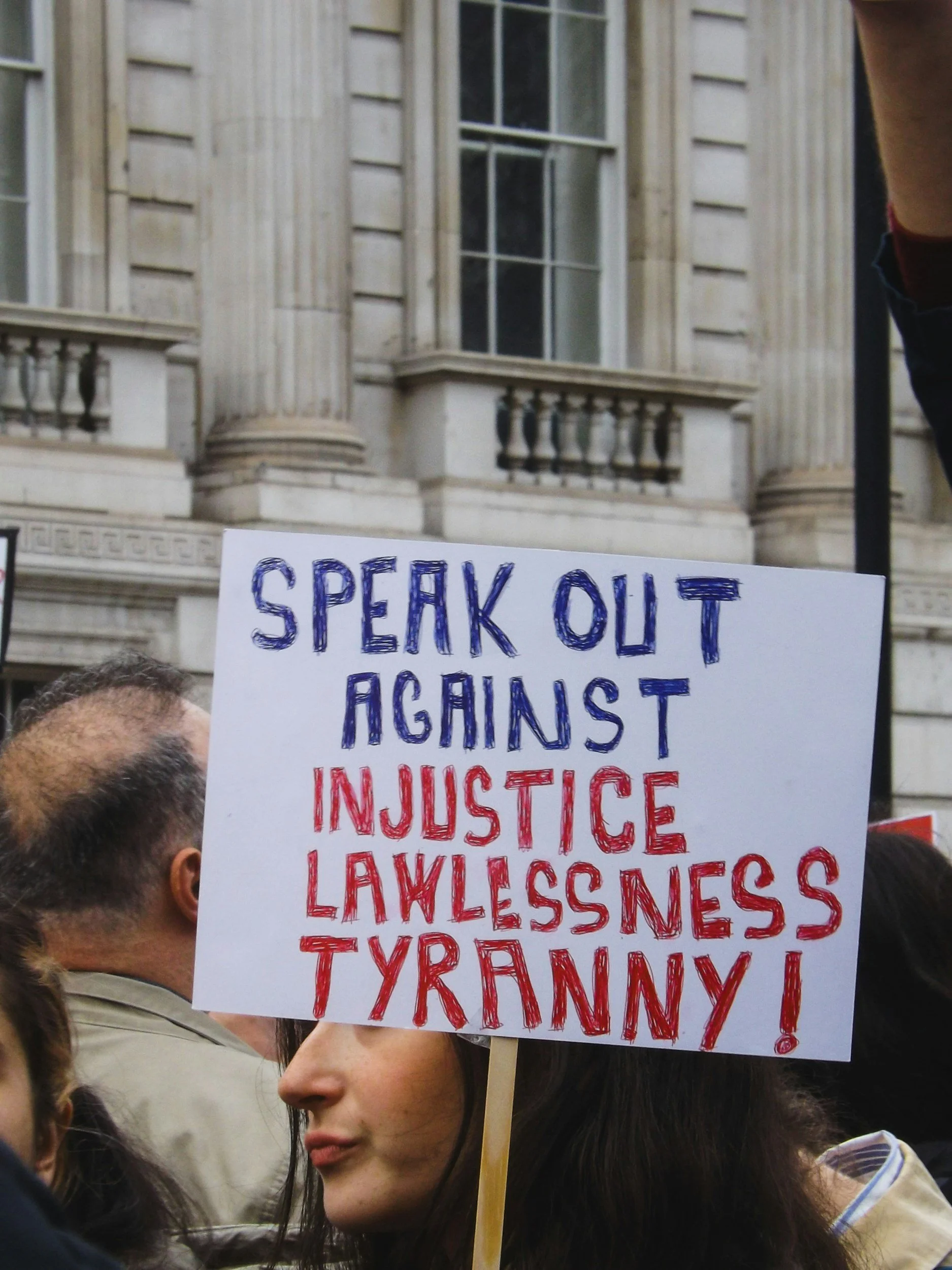 A person holding a sign at a protest, with a building with columns in the background. The sign reads 'Speak Out Against Injustice, Lawlessness, Tyranny!' in red and blue letters.