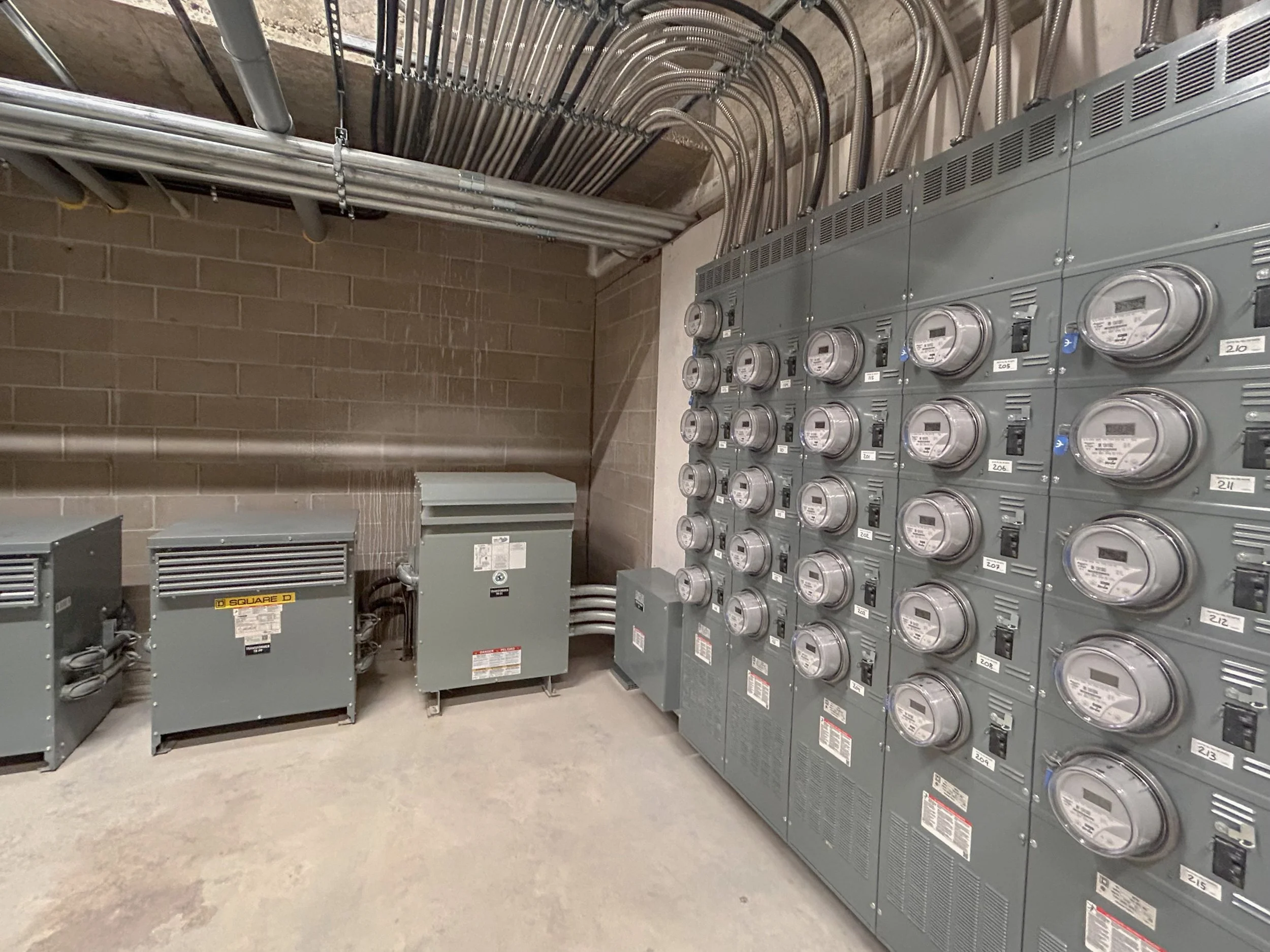 Utility room with multiple electrical meters and conduits mounted on the wall.