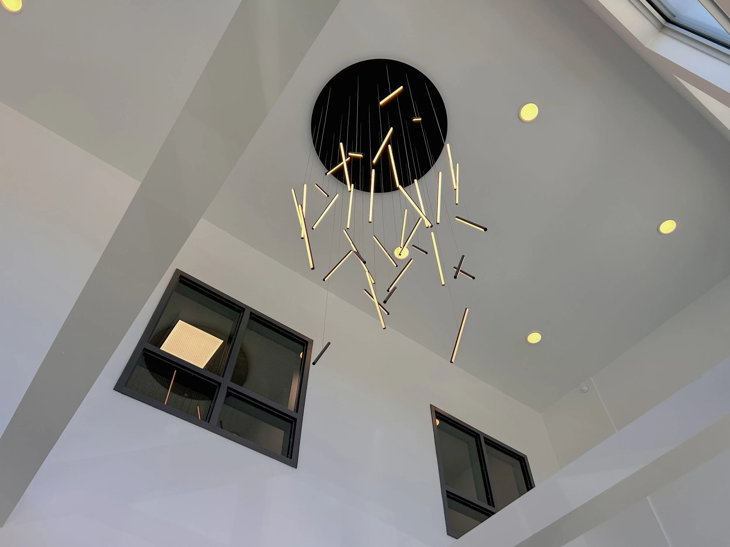 Modern chandelier with elongated yellow and black light fixtures hanging from a black circular base on a white ceiling with ceiling lights and windows.