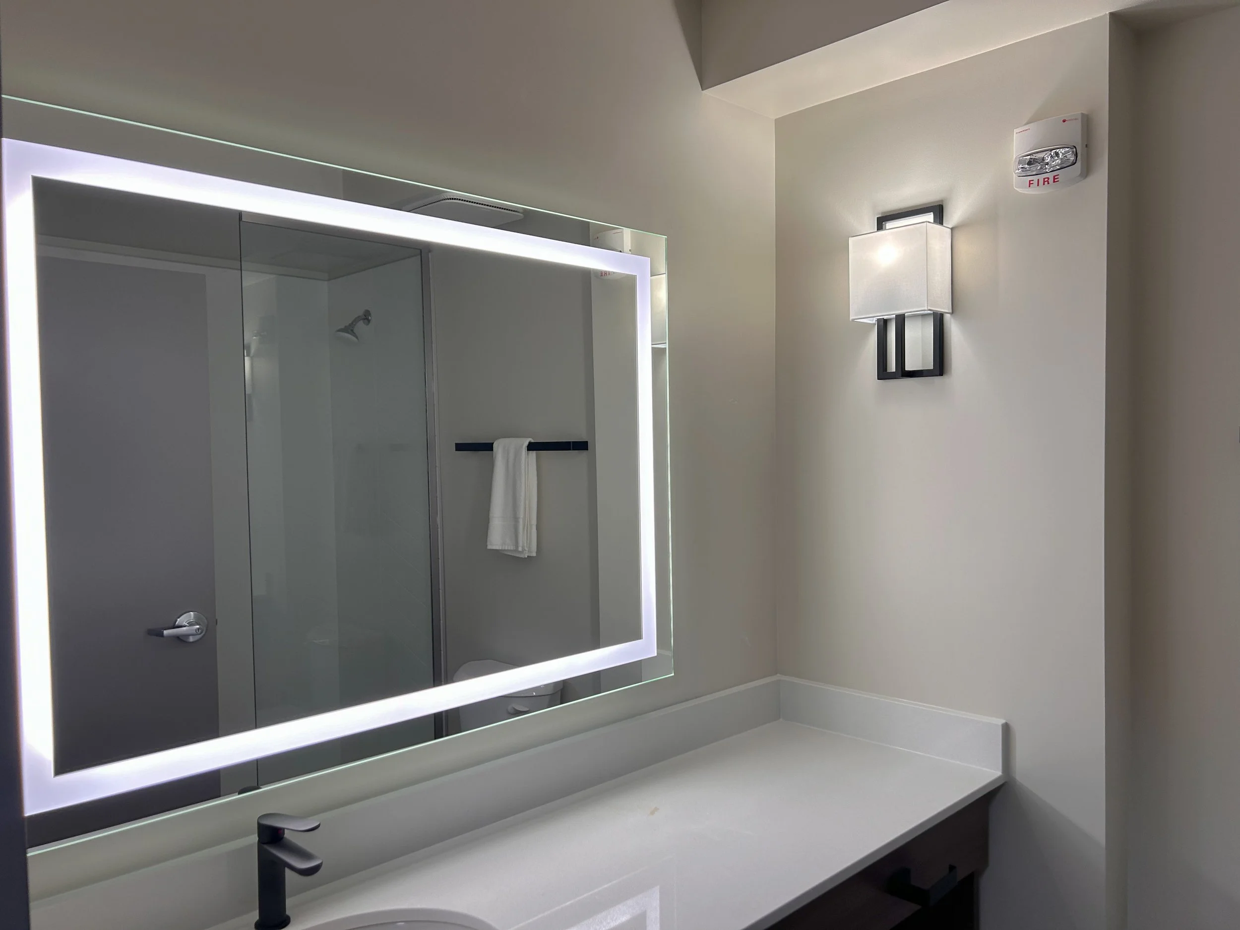 A bathroom with a large illuminated mirror, a light-colored countertop, a black faucet, a towel hanging on a black bar, a door to a shower or toilet area, a wall-mounted light fixture, and a fire alarm on the wall.