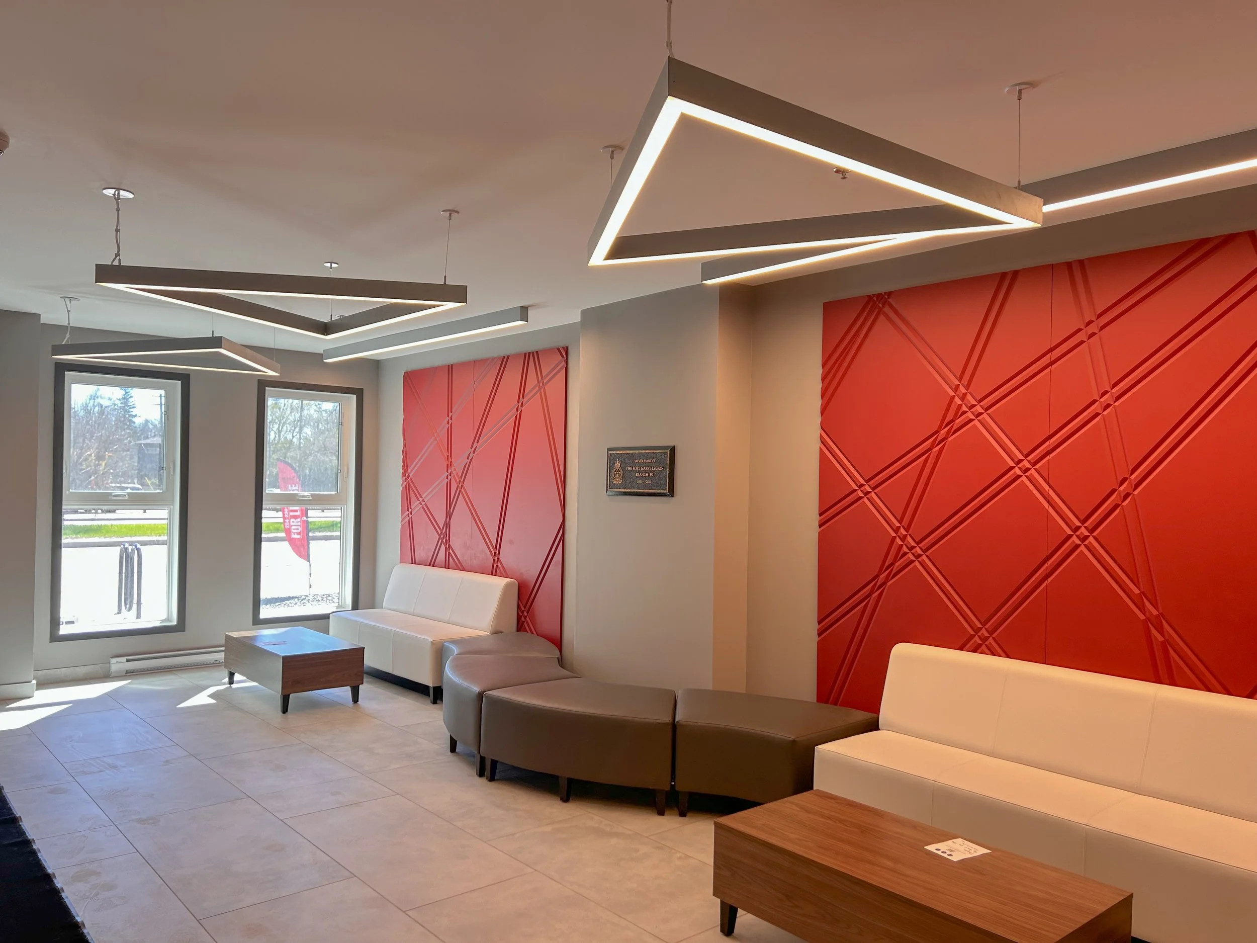 Modern hotel lobby with white and beige couches, a wooden coffee table, large windows, red accent wall panels, and contemporary geometric ceiling lights.