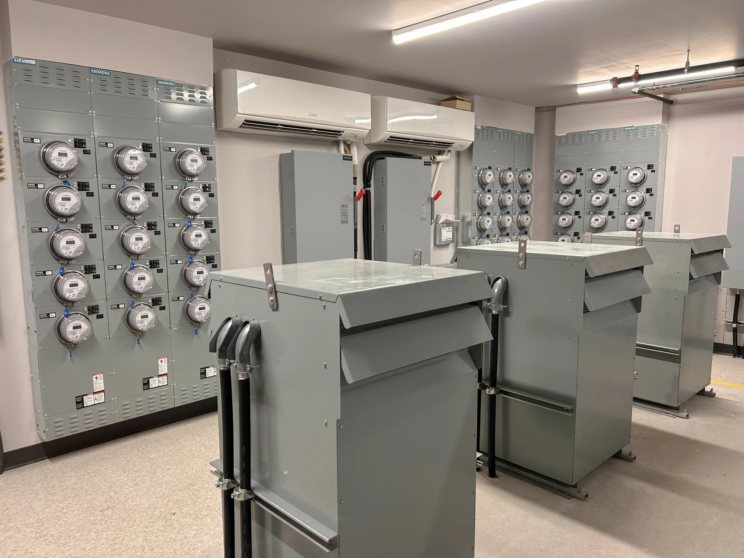 Electrical room with multiple electric meters, air conditioning units, and electrical panels.