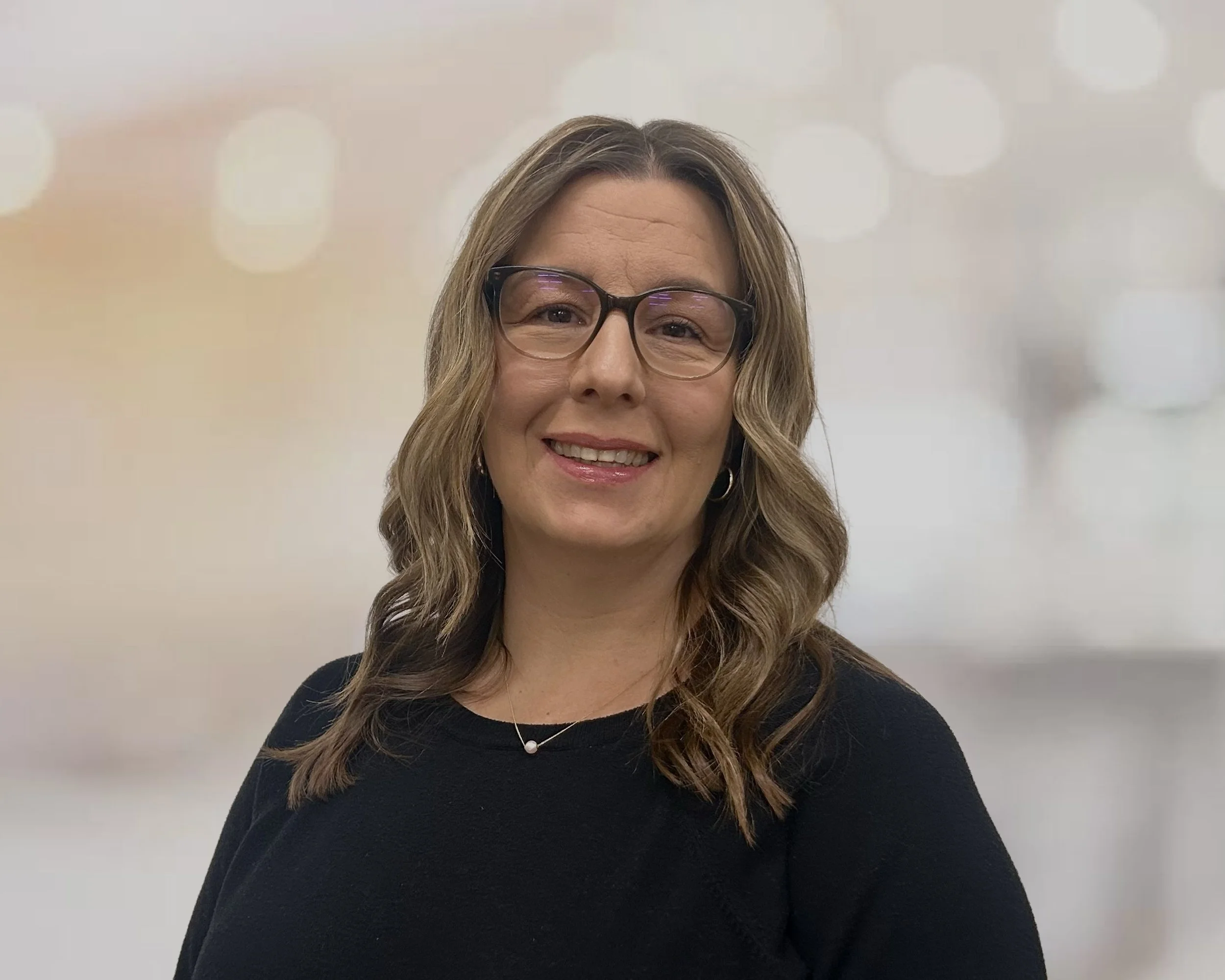 A man with dark, curly hair and glasses looking to the side in a light-colored room with a plant in the background.