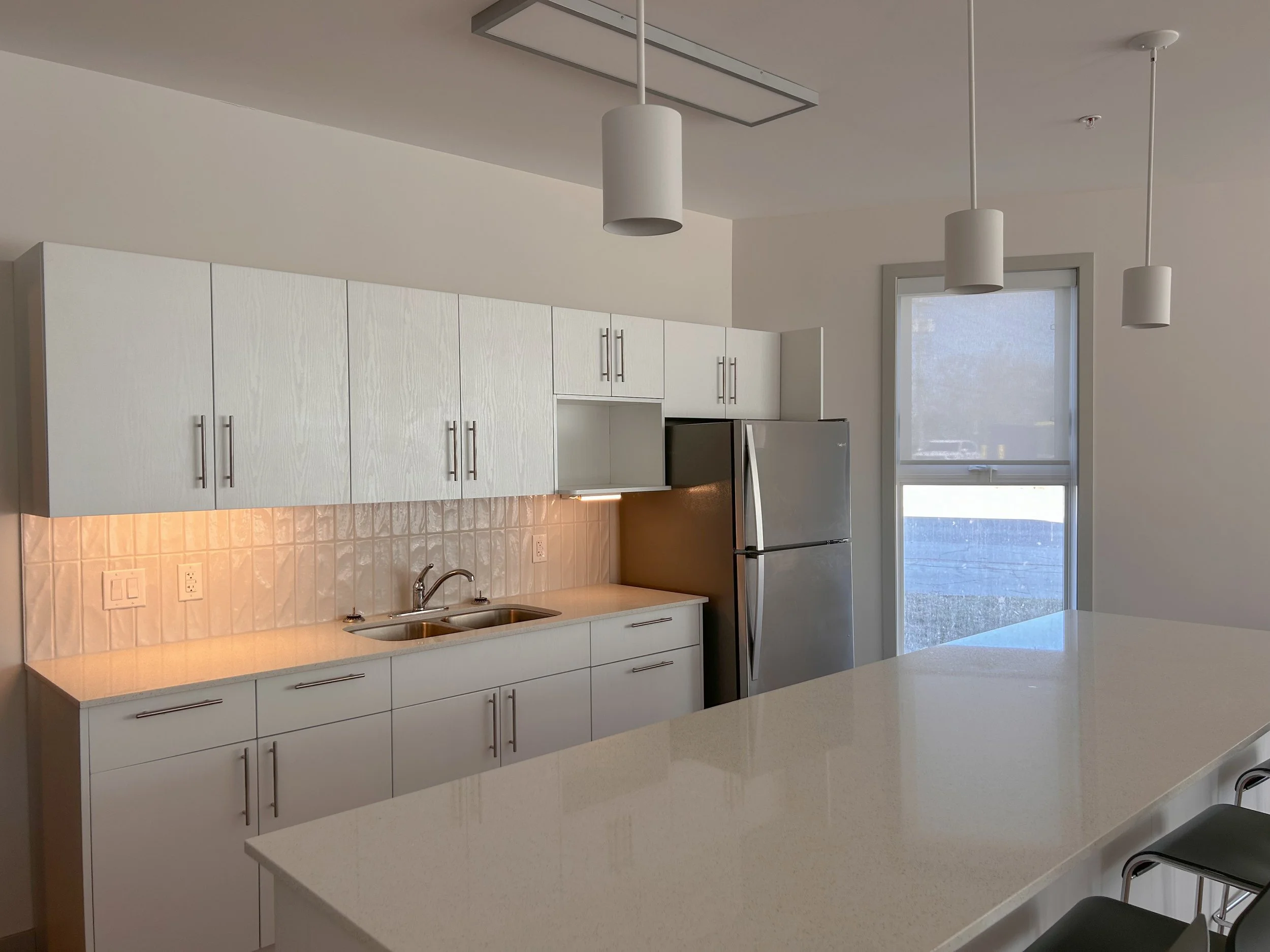 Modern kitchen with white cabinets, stainless steel refrigerator, double sink, beige tile backsplash, large window, and white island counter with black chairs.