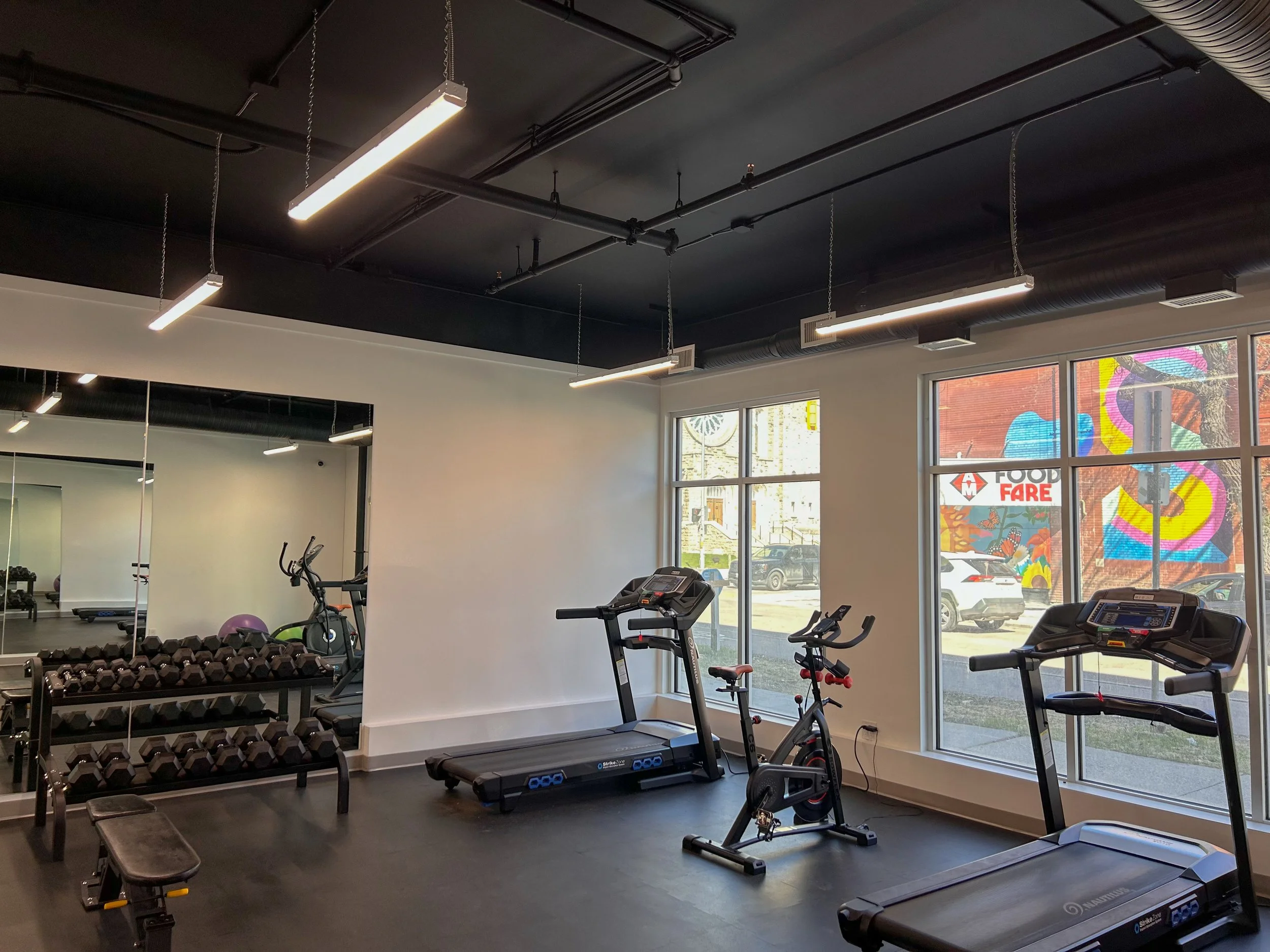Indoor gym with exercise equipment including a treadmill, stationary bike, and dumbbells, with natural light coming through large windows.