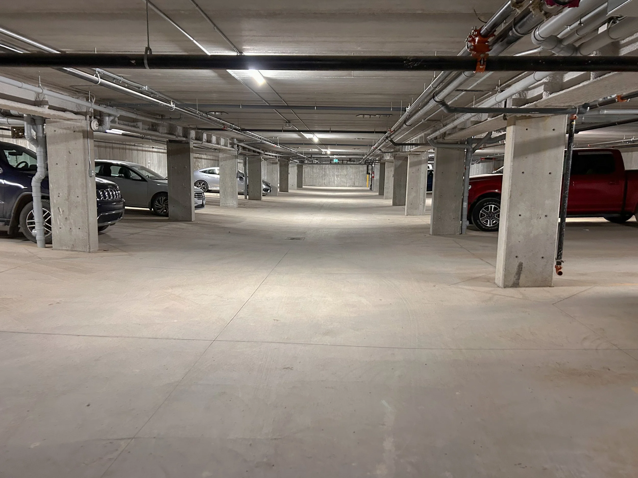 Empty indoor parking garage with parked cars on both sides, concrete pillars, and overhead pipes.
