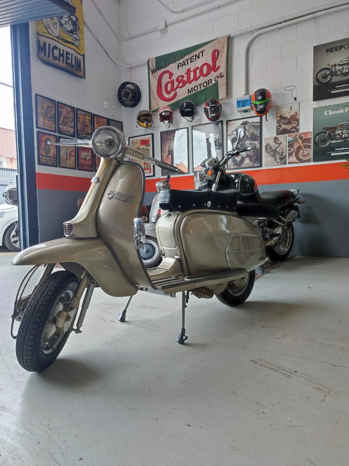 A vintage beige Vespa scooter parked on a garage floor with a black motorcycle behind it. The garage has posters, helmets, and memorabilia on the wall, including an orange and white Castrol sign. Natural light coming from the opening on the left.