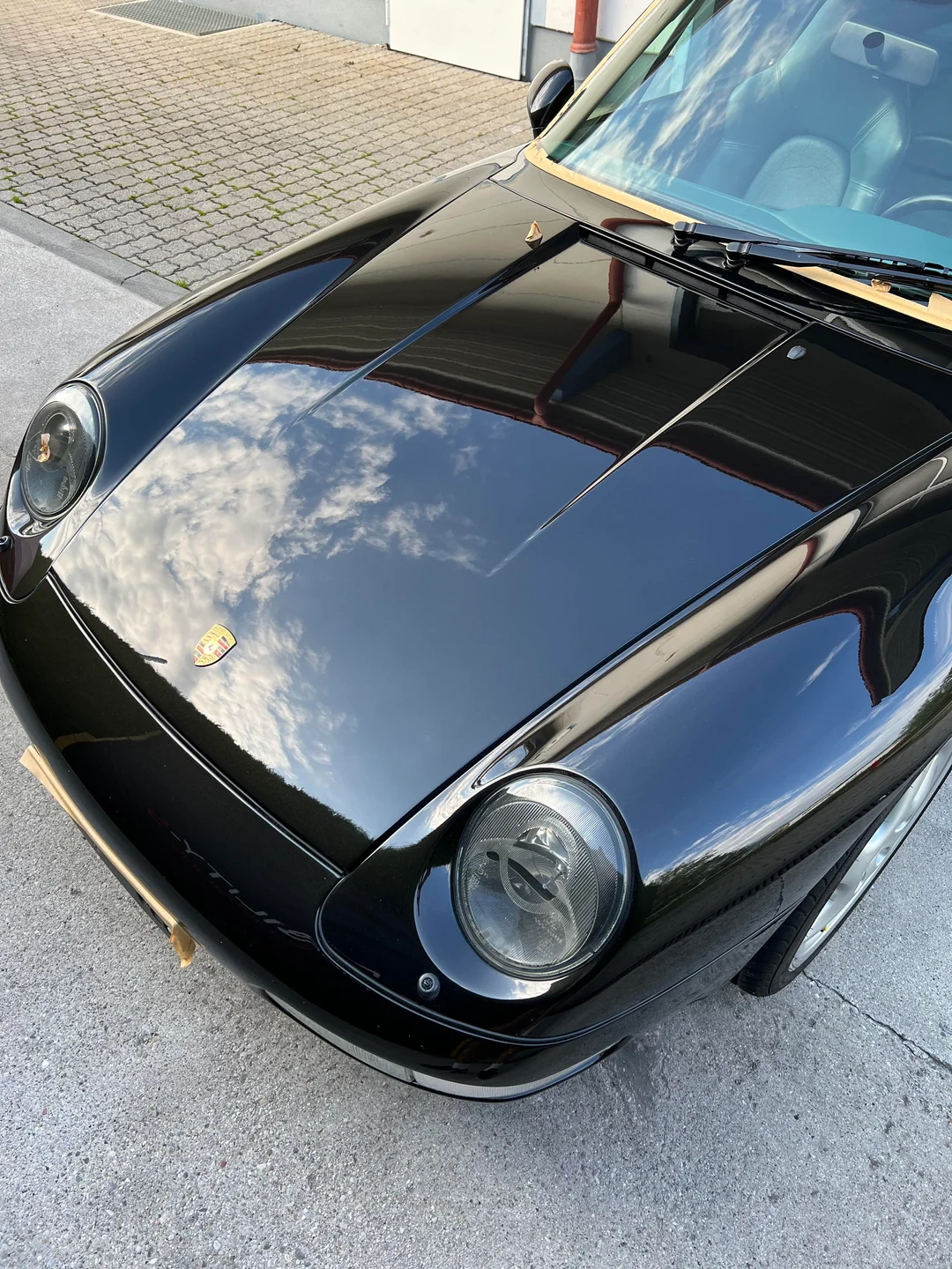 Auto deportivo Porsche clásico negro estacionado en el suelo, reflejando el cielo con nubes en el capó y el parabrisas.