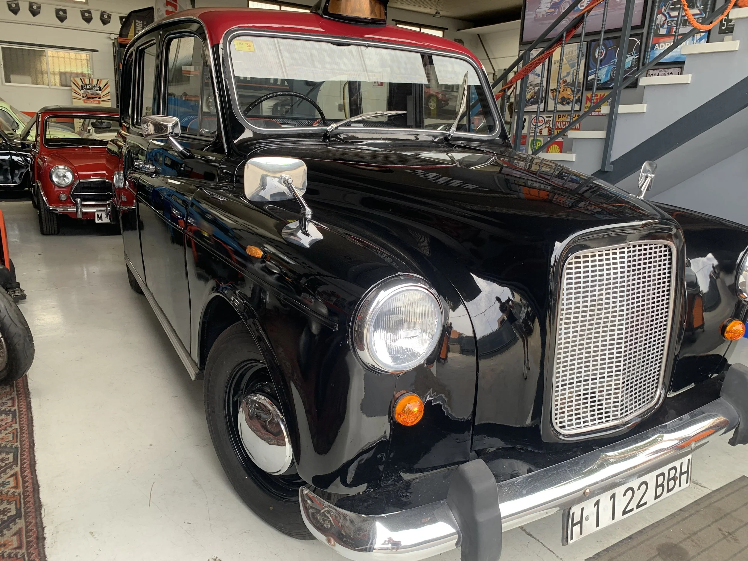 Coche clásico negro con parrilla metálica, espejos laterales y ruedas con cubiertas, exhibido en un espacio interior con otros autos antiguos en el fondo.