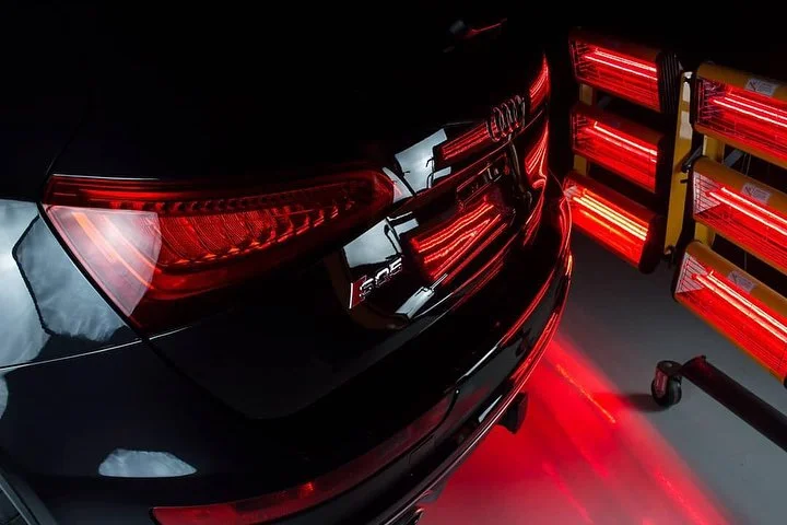 La parte trasera de un coche negro con luces LED rojas encendidas y un equipo de calentadores de cerámica rojos y amarillos en un taller.