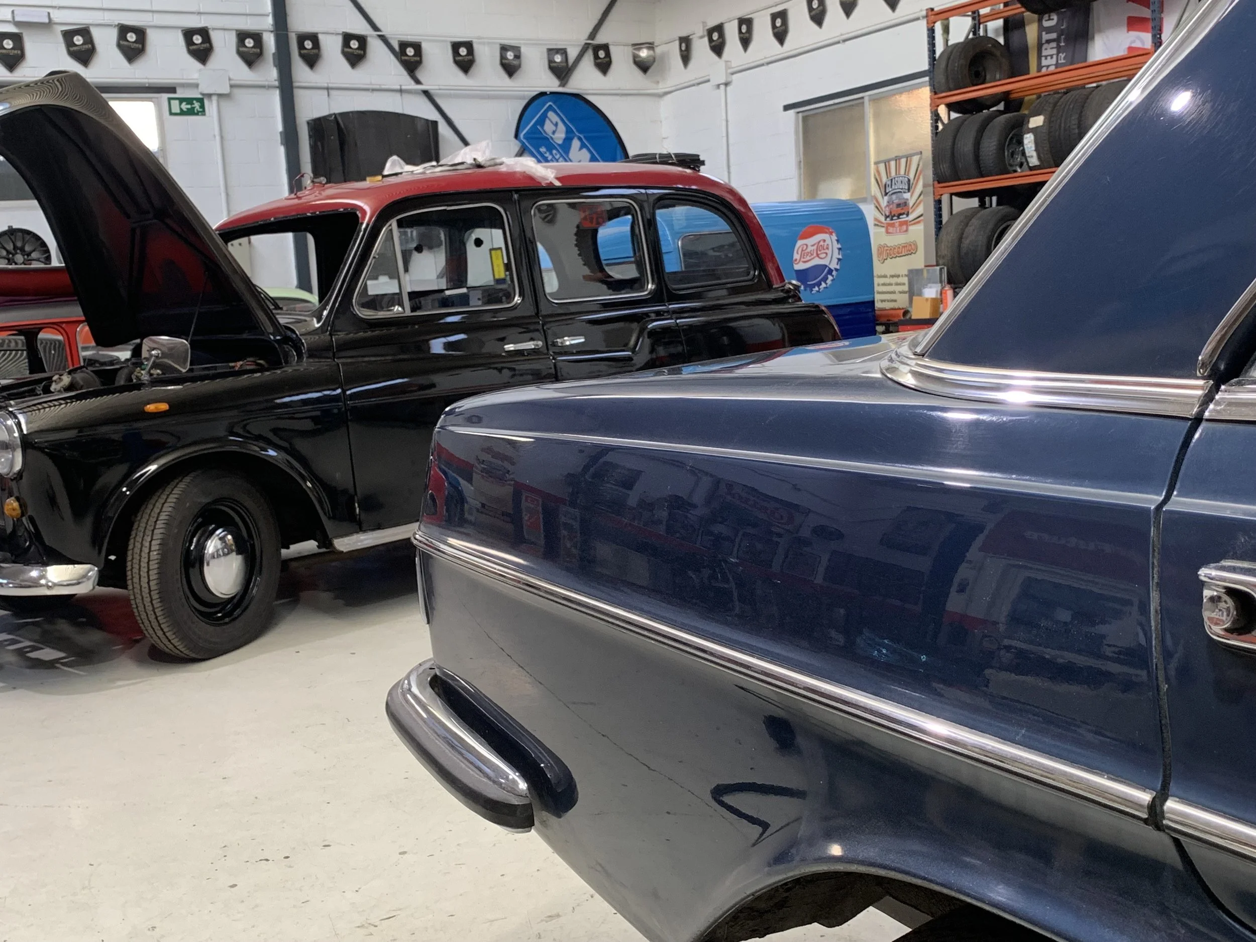Vintage cars parked inside a garage with tires stored on shelves and banners hanging from the ceiling.