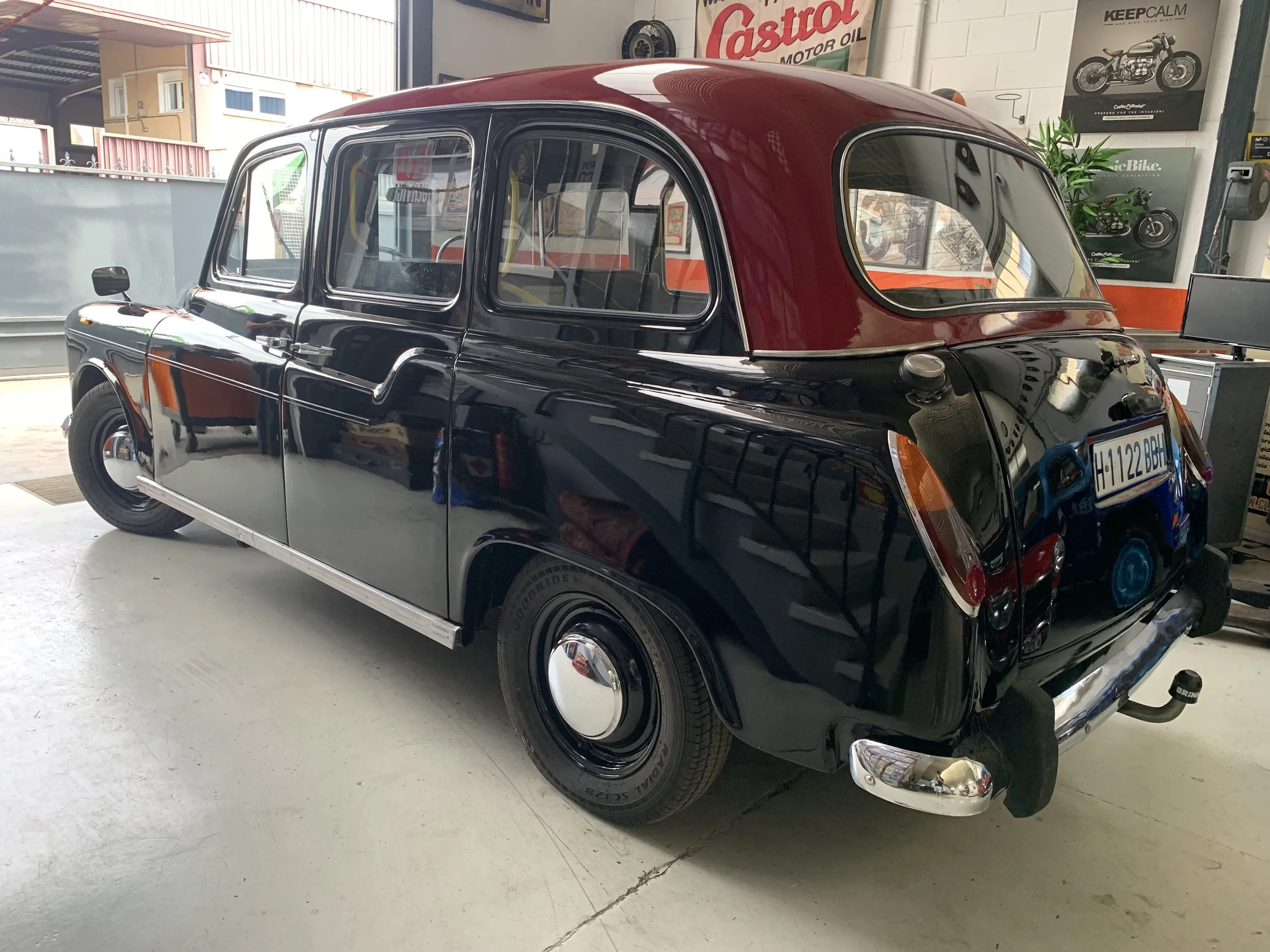 Coche clásico negro con techo rojo en exhibición interior, en un taller o museo de autos, con posters de motocicletas en la pared y un monitor en el fondo.