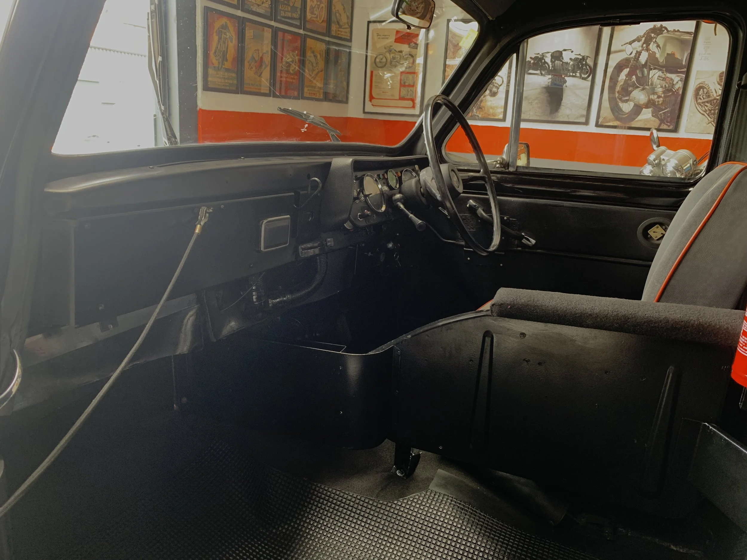 Interior de un vehículo clásico o antiguo con puesto de conductor, tablero, volante y ventanas, decorado con posters y motocicletas en exhibición.