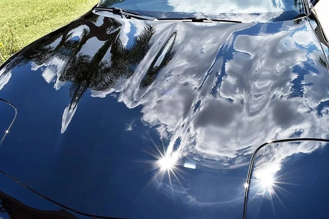 Capó de coche negro reflejando el cielo con nubes y árboles.