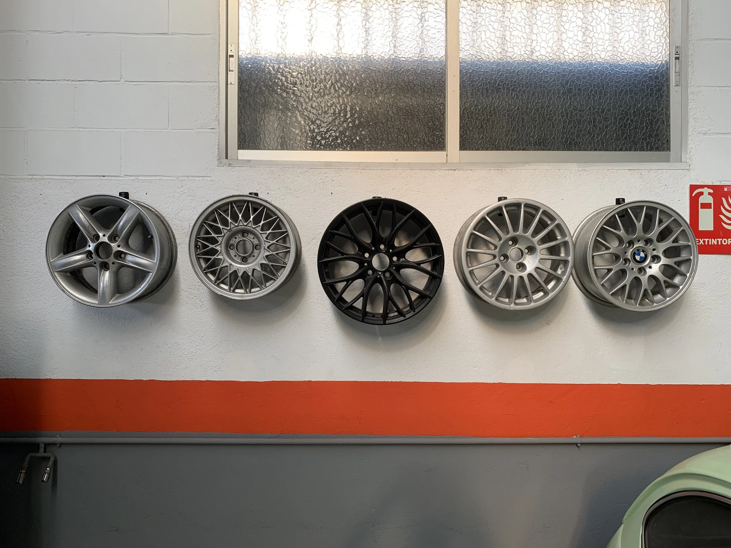 Five car wheels mounted on a white wall above an orange stripe, with a window above and a red fire extinguisher sign on the right.