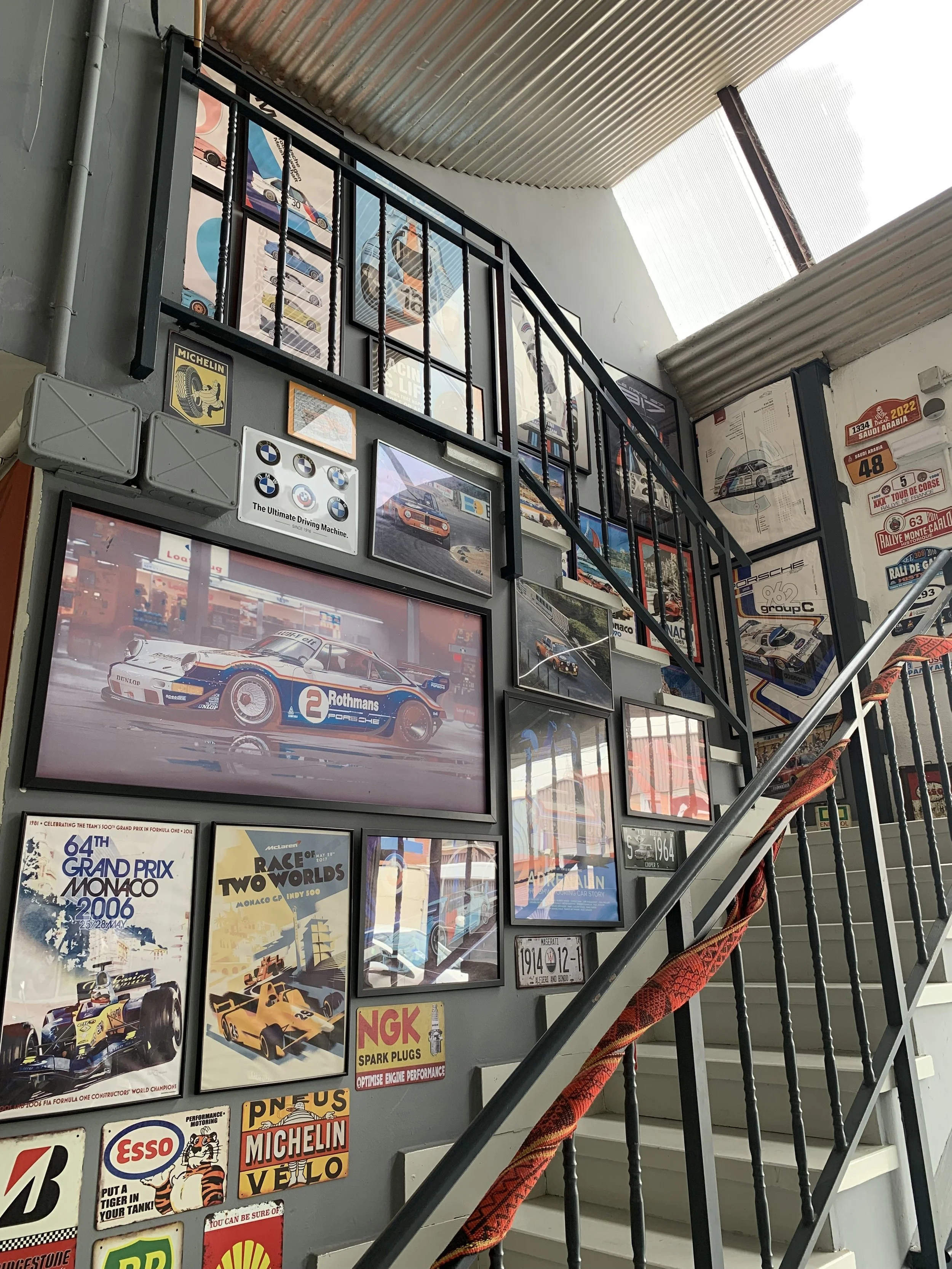 A staircase wall decorated with various vintage car posters, photographs, and license plates, featuring racing cars and racing events.