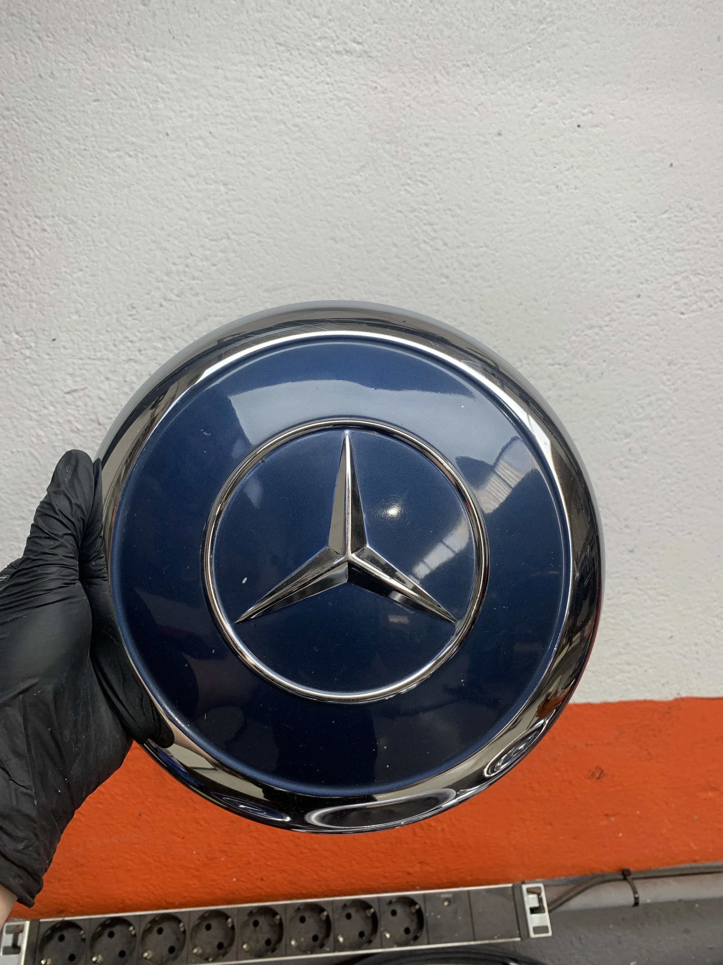 A person wearing a black glove holding a round Mercedes-Benz hubcap with a blue background and the Mercedes-Benz logo in the center.