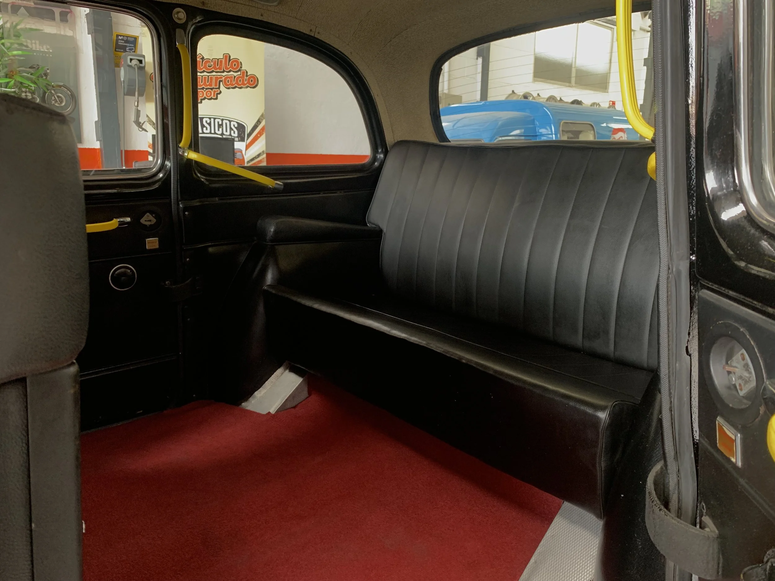 Interior de un autobús antiguo con asiento de vinilo negro, ventanas pequeñas, pasamanos amarillos y alfombra roja en el suelo.