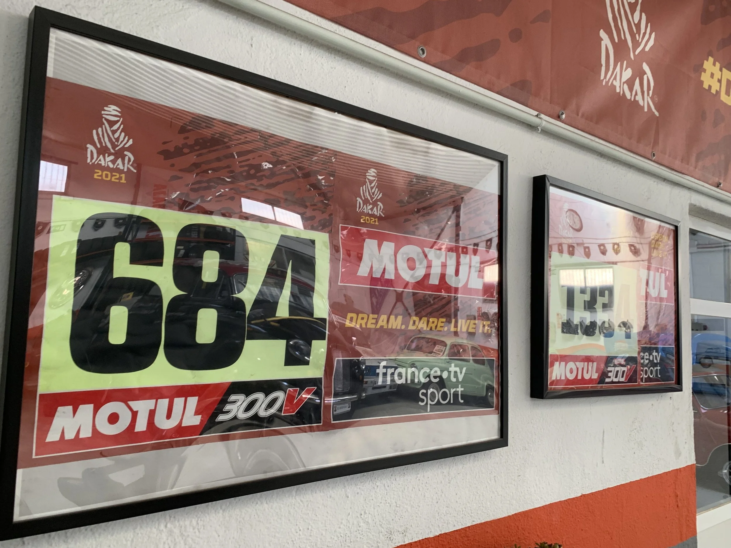 Framed posters on a wall promoting the 2021 Dakar Rally with the number 684, Motul, and France.tv sport logos, and the slogan 'Dream. Dare. Live it.'