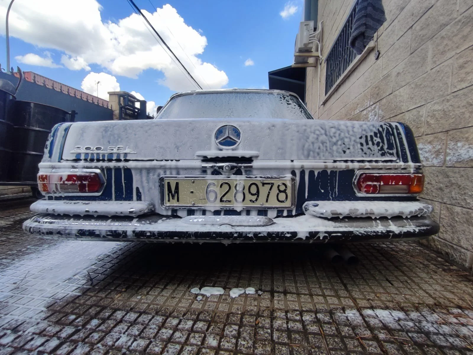 Rear view of a white vintage Mercedes-Benz 200 SEL car covered in soap suds during a car wash, parked on a stone-textured driveway beside a brick wall and a black trash bin with a cloudy sky above.
