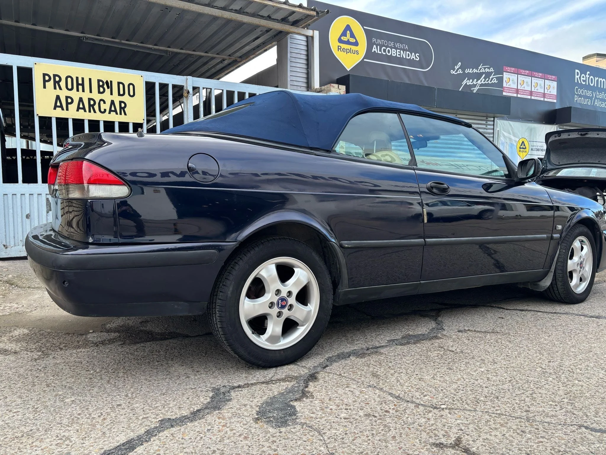 Black convertible car with a soft top parked outside a building with signs in Spanish, and an open hood on the front passenger side.