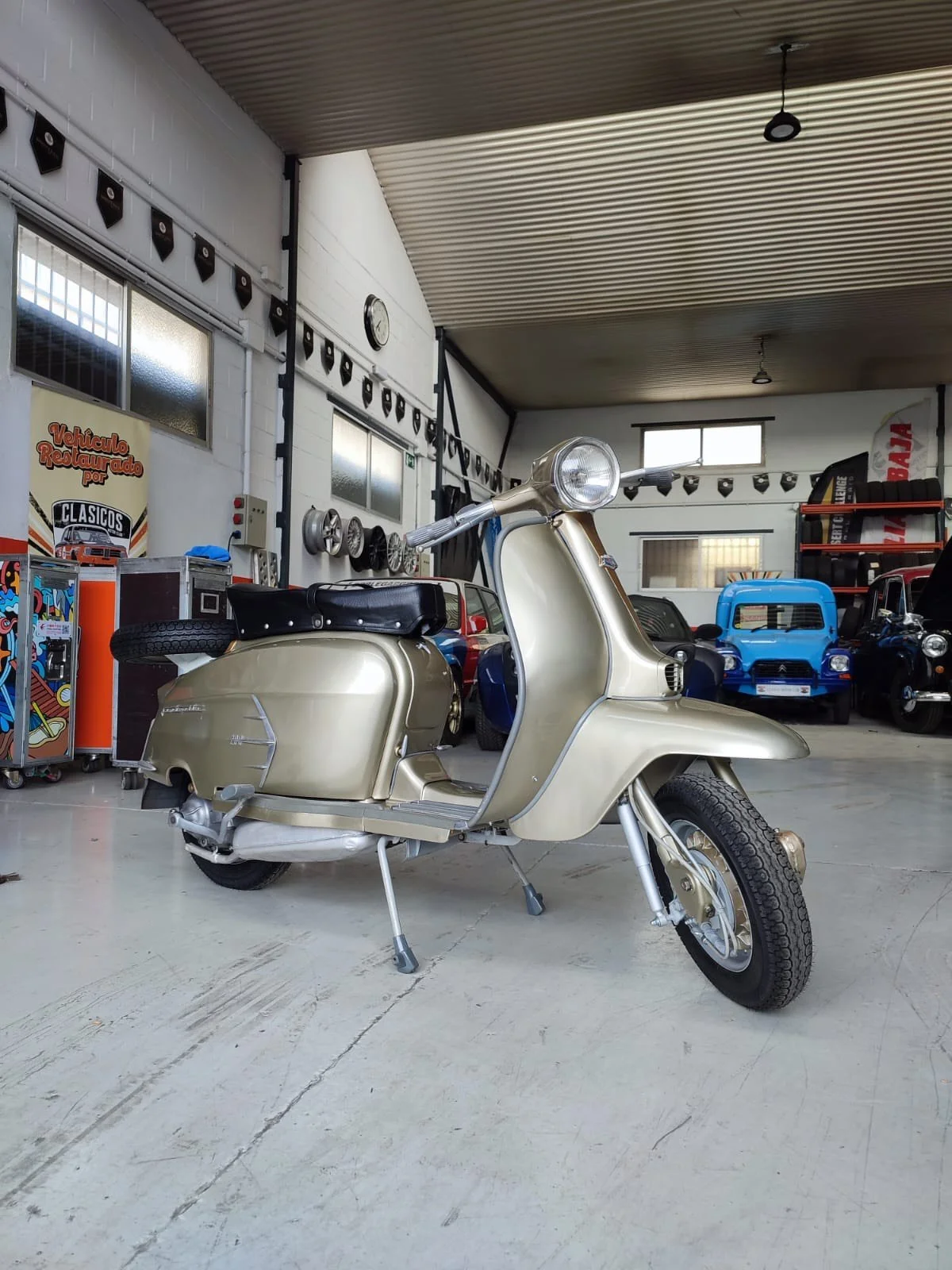 Vintage cream-colored scooter parked inside a garage with classic cars in the background.