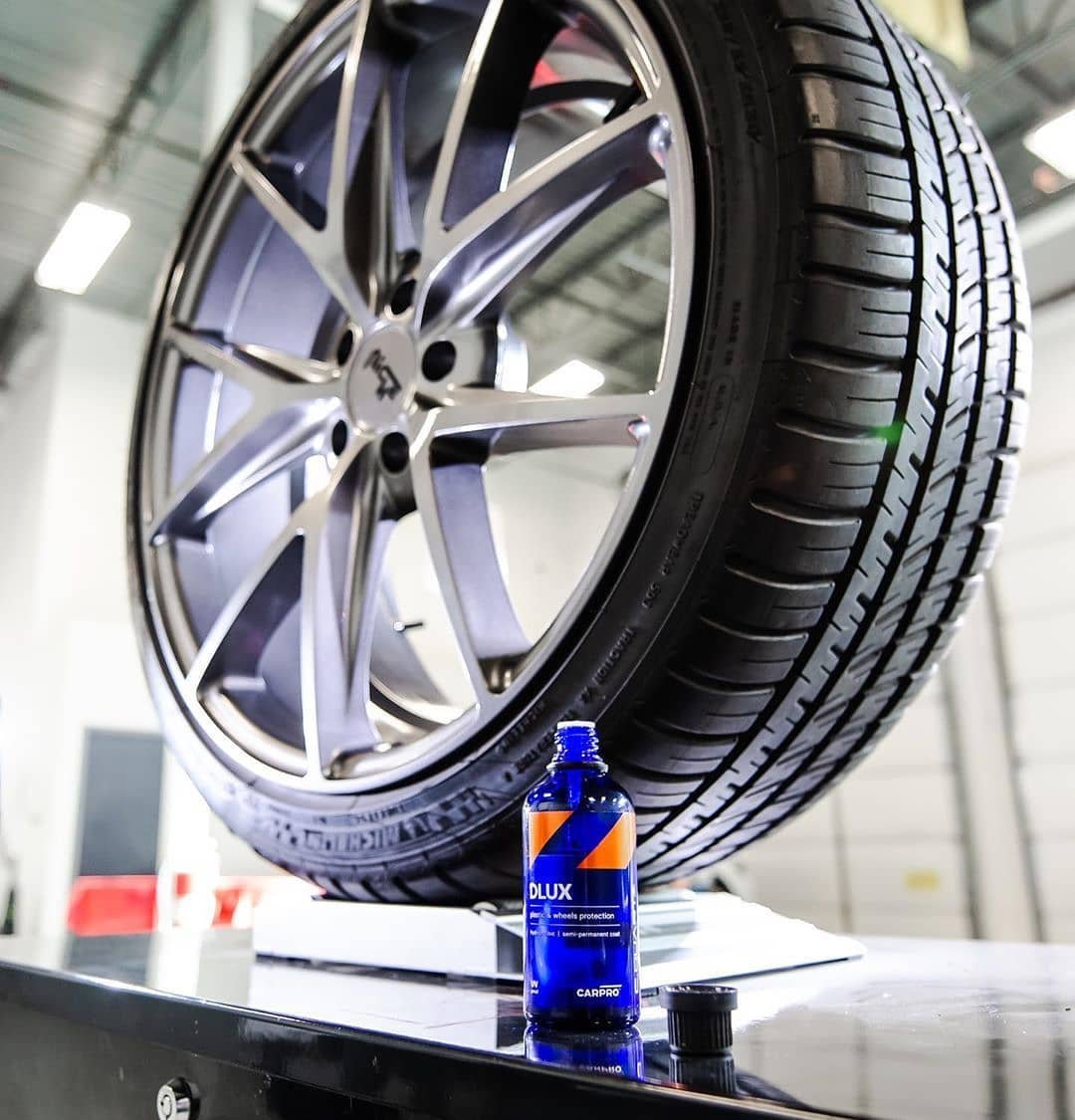 A close-up of a car tire mounted on a shiny alloy wheel, with a bottle of car wheel protection spray in the foreground on a workbench inside a garage or workshop.