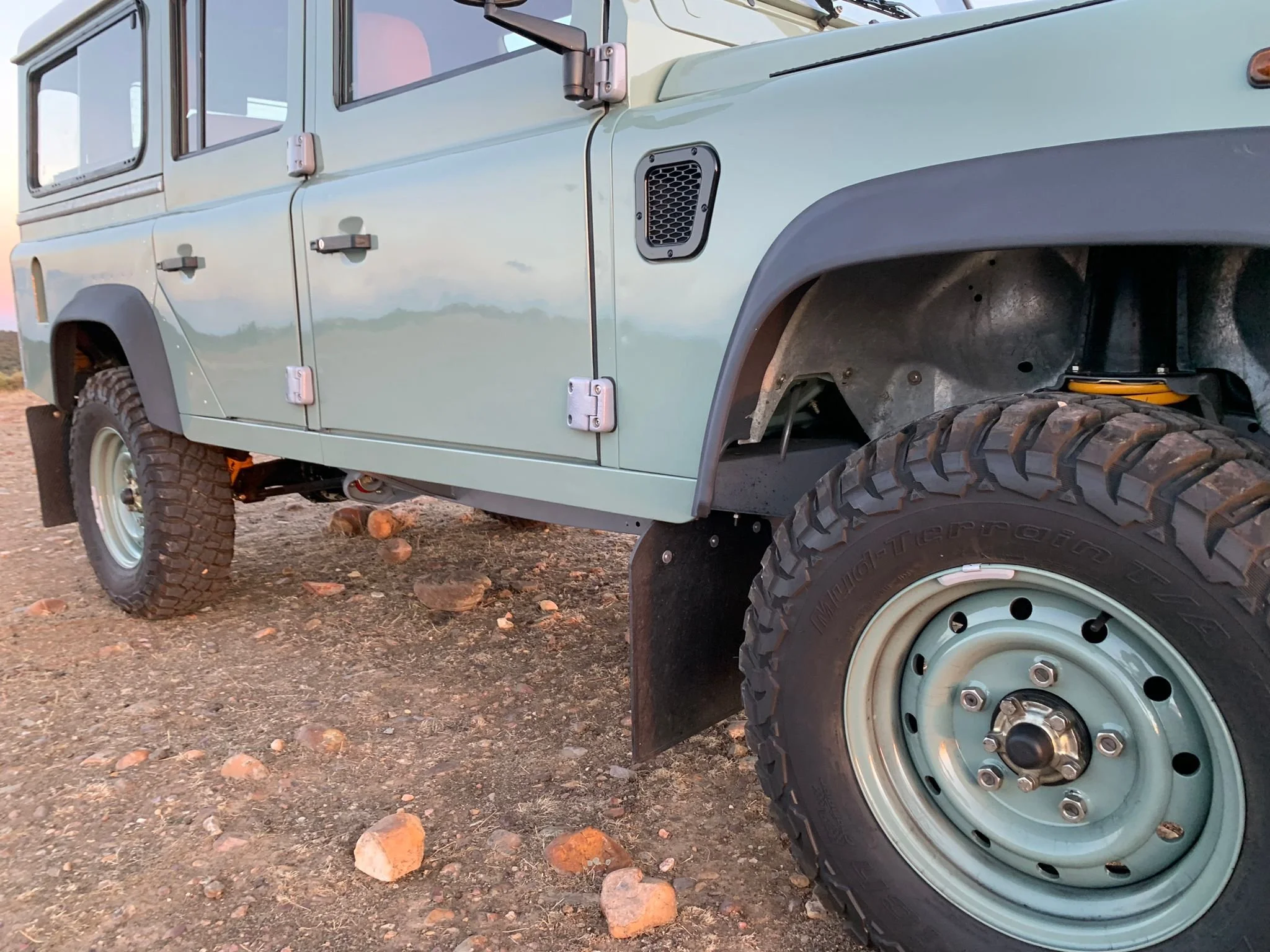 Vehículo todoterreno de color verde claro en un terreno con piedras, mostrando la rueda delantera y parte del costado del automóvil.