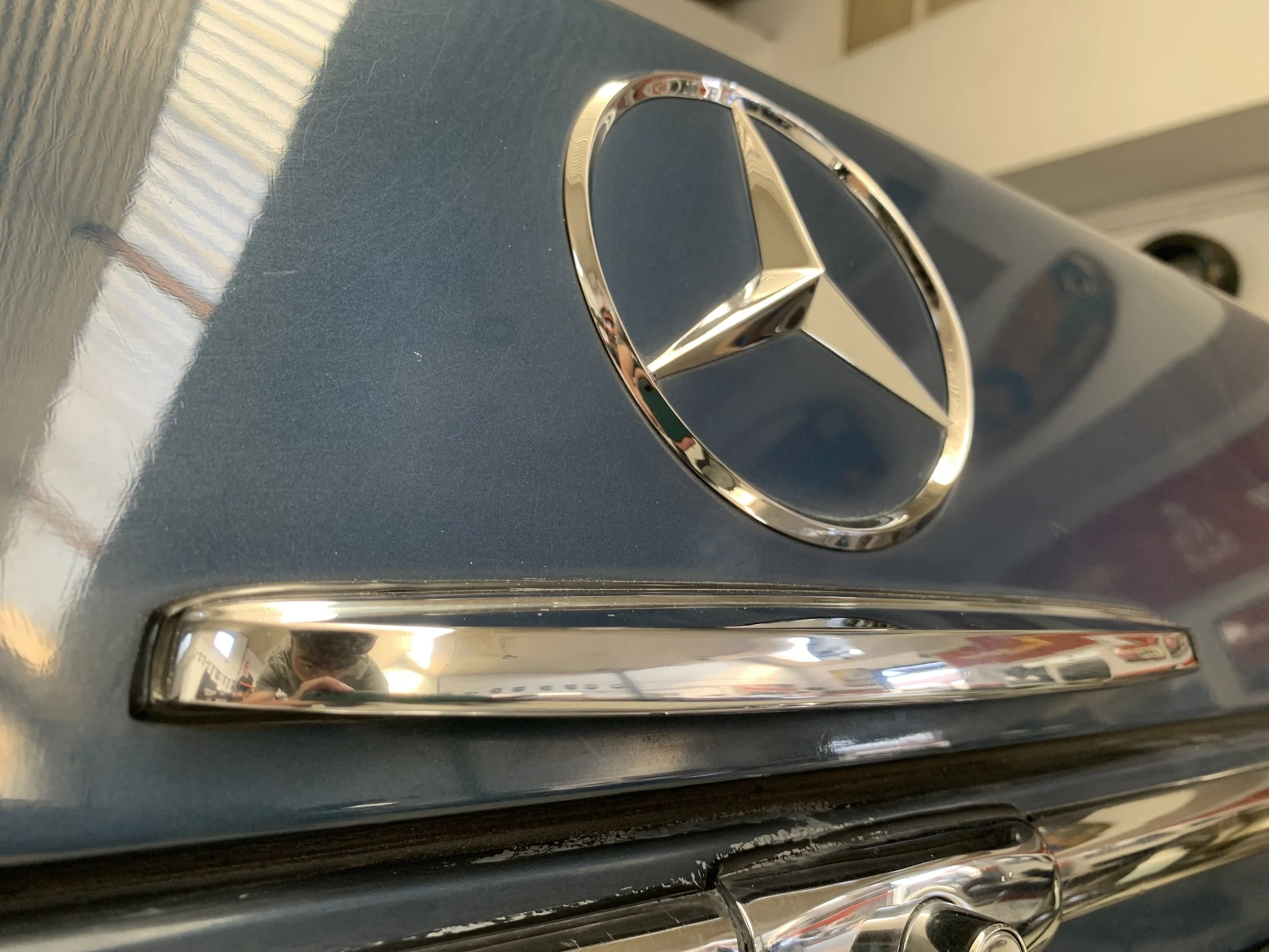Close-up of a metallic Mercedes-Benz emblem on a dark gray car hood, with a chrome strip and a reflection of the person taking the photo.