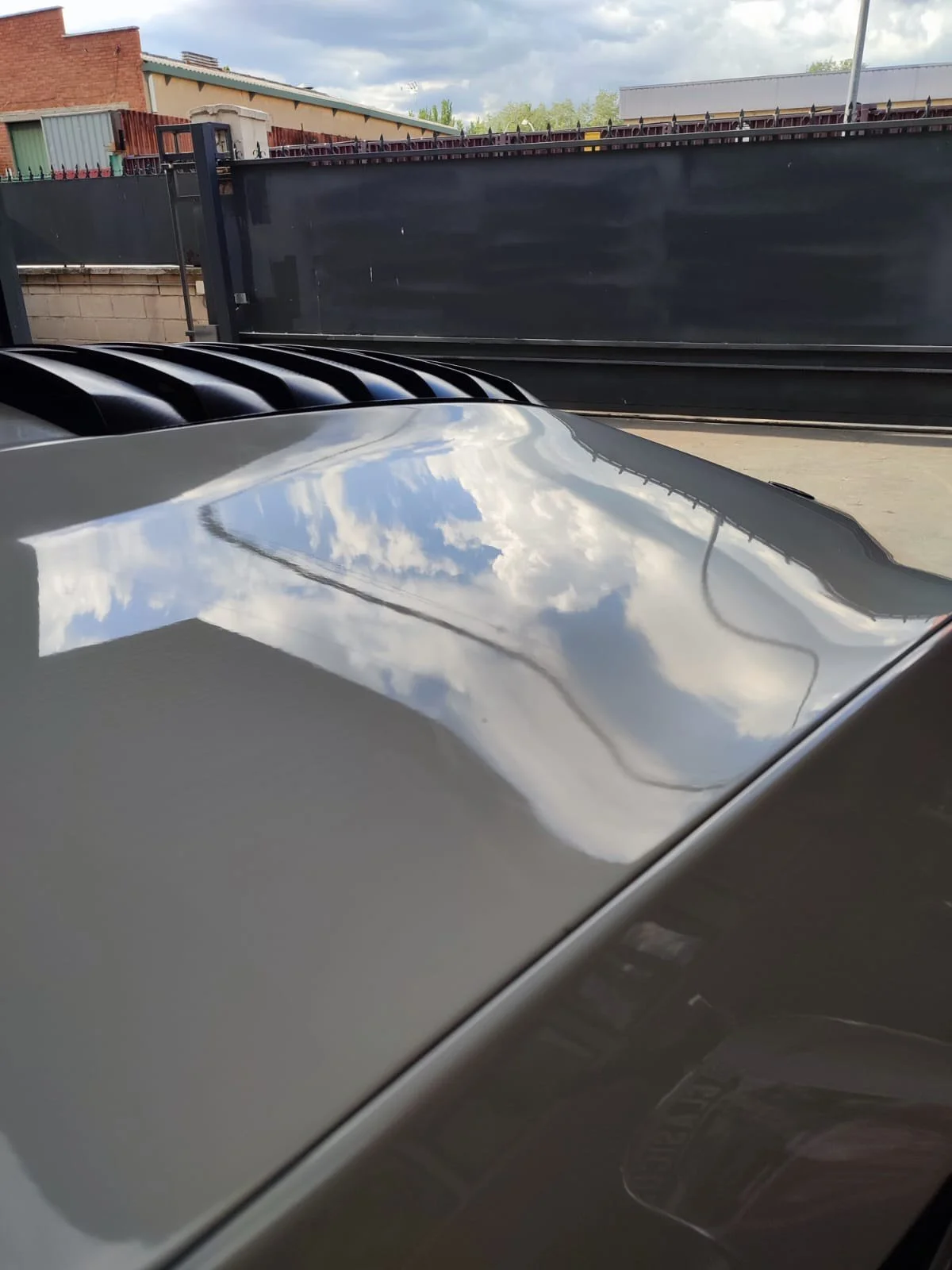 Reflection of clouds in the shiny hood of a car parked outside near a black gate.