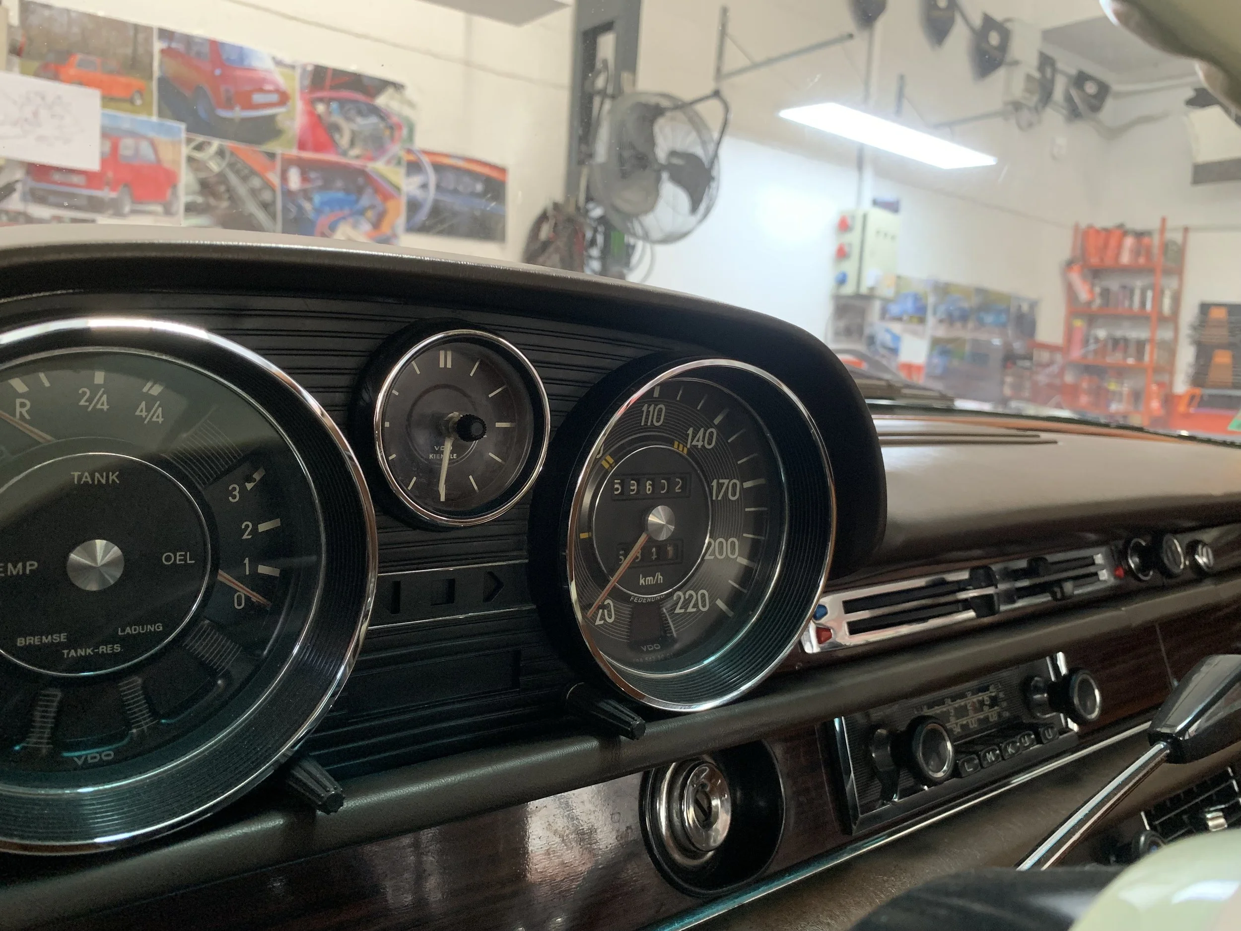Dashboard of a vintage car showing speedometer, fuel gauge, and other instrument panels inside a garage with shelves and posters in the background.