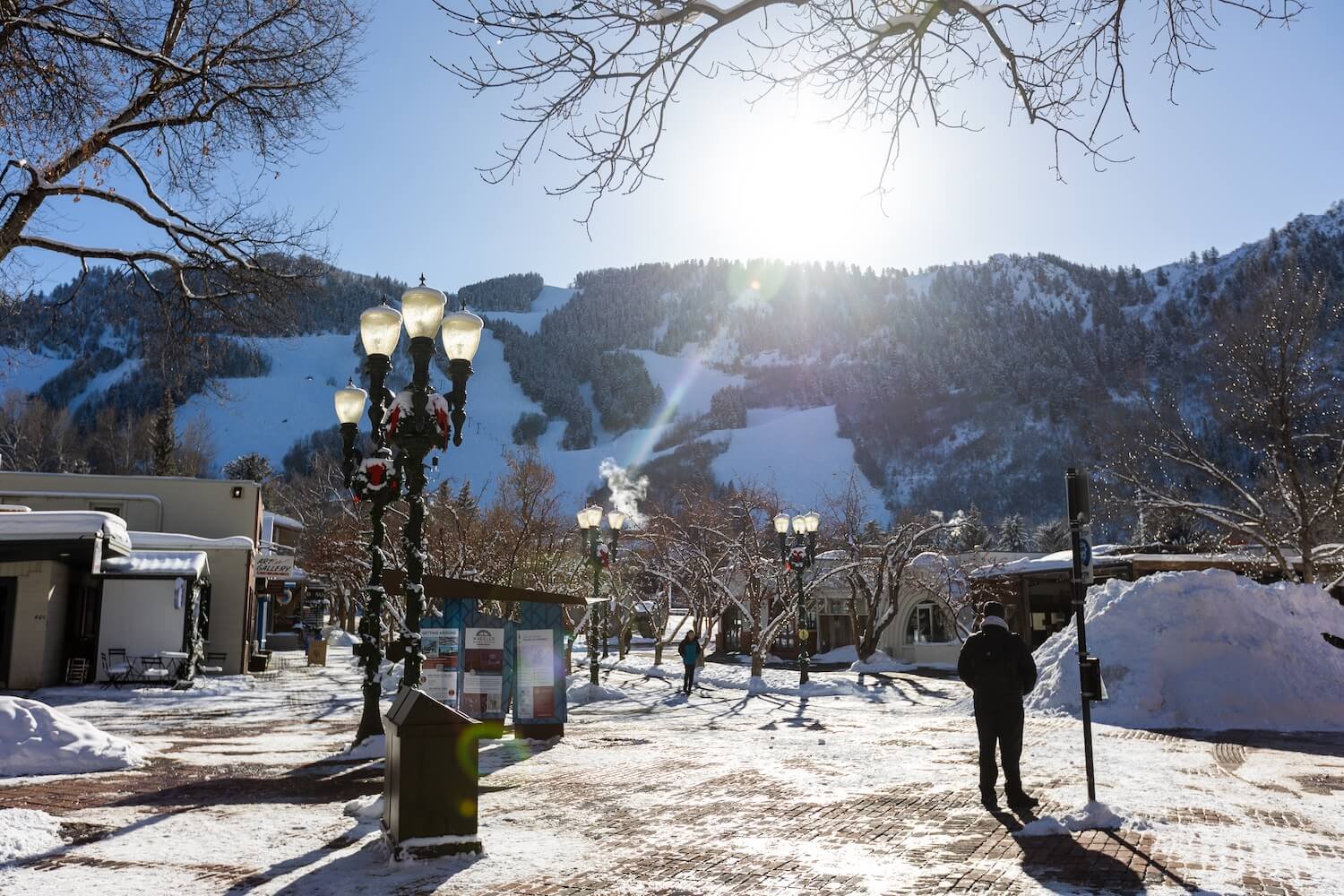 A snowy mountain town square with street lamps decorated for the holidays, snow-covered trees, and people walking on a cleared path. The sun shines brightly over the mountains in the background.