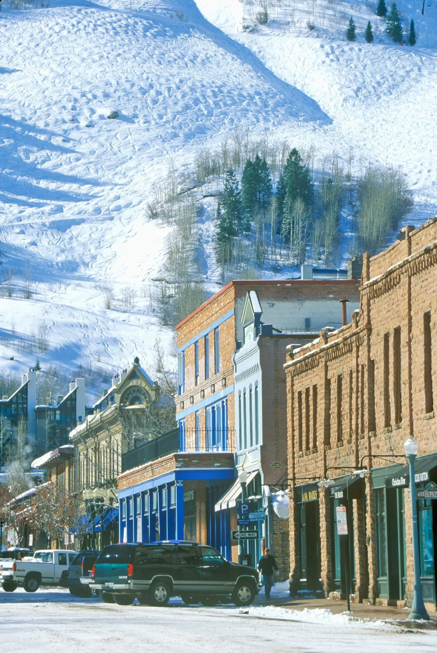 Snow-covered buildings and street in a mountain town with a snowy hillside and trees in the background.