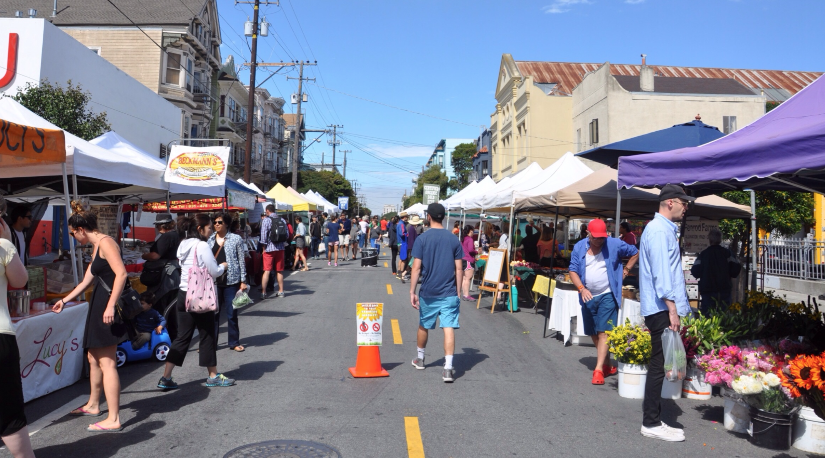 People shopping at a street market with vendor stalls and tents on both sides, under a bright blue sky.