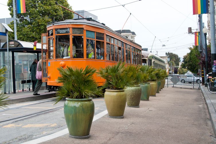 Orange vintage trolley on street with large potted plants in front, pedestrians nearby, and city buildings in background.