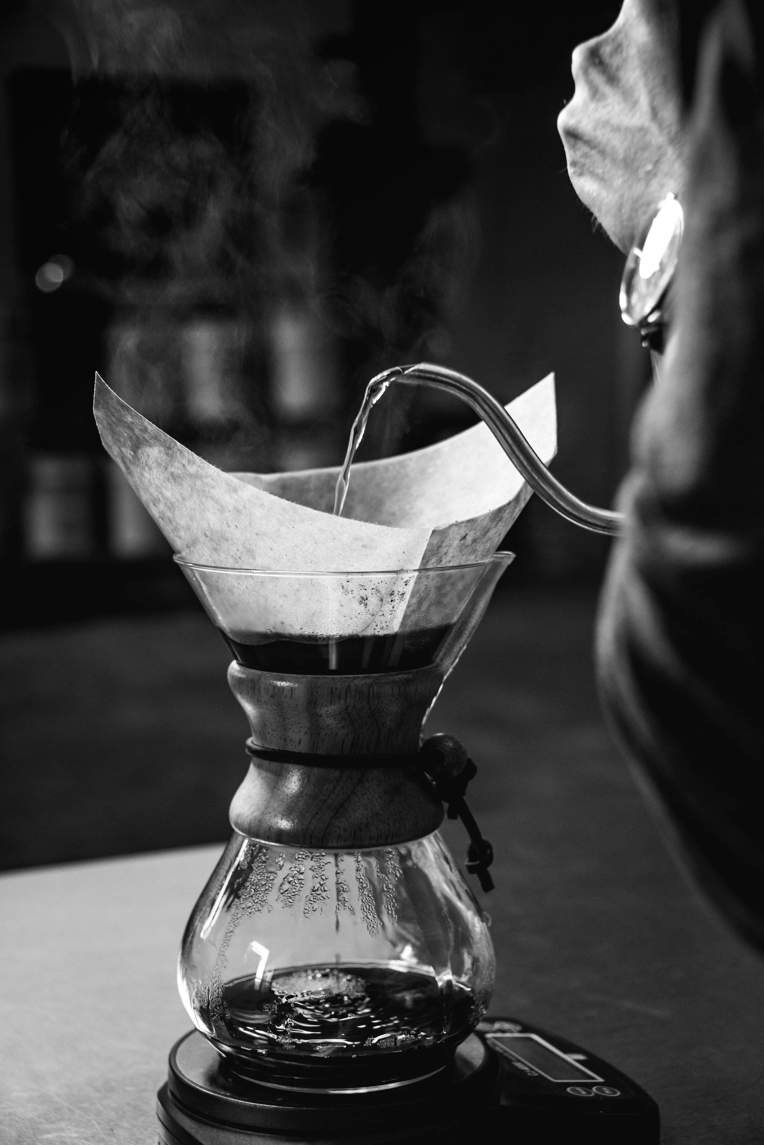 Black and white photo of a coffee brewing process using a pour-over method, with steam rising from the hot coffee in a glass vessel, a paper filter, and a metal gooseneck kettle pouring hot water.
