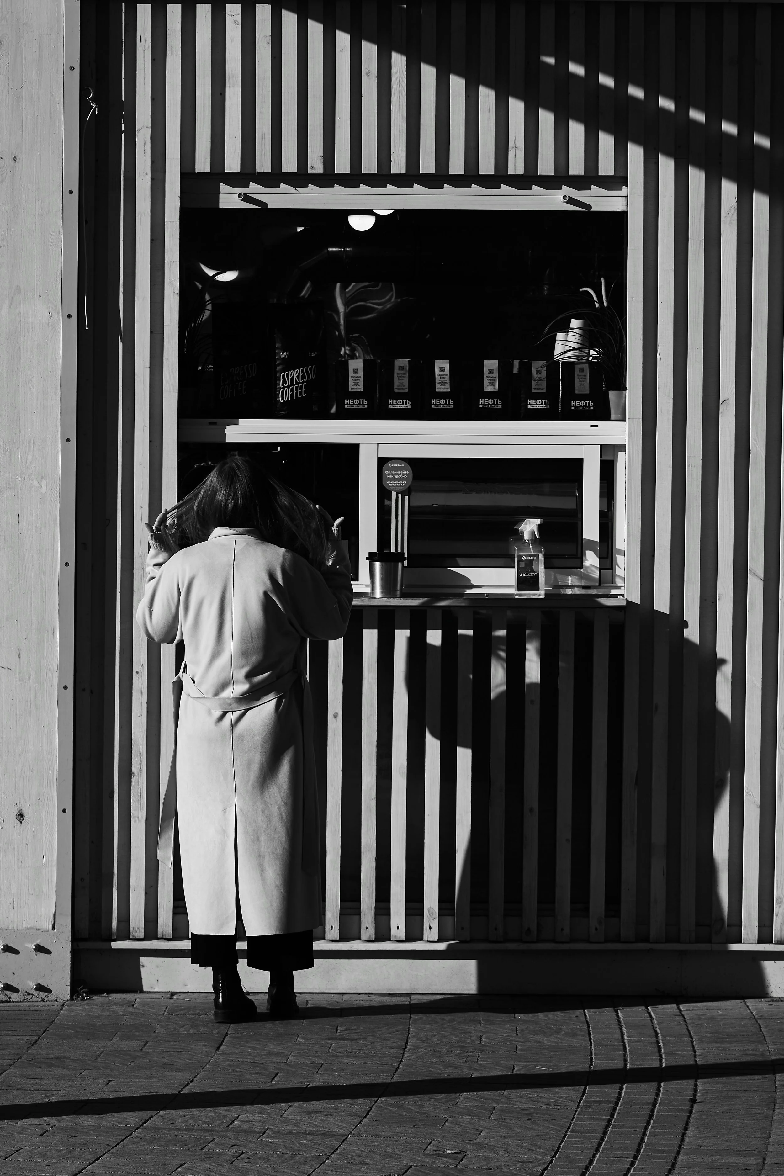 A woman in a long coat is standing at a small window of a wooden building, appearing to place an order for coffee, with sunlight casting shadows on the scene.